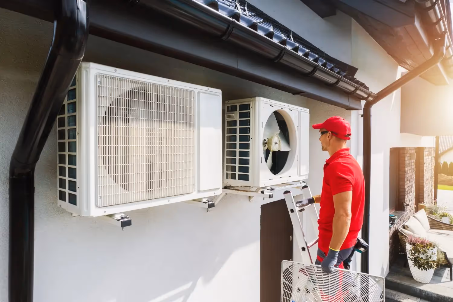 A technician in a red uniform stands on a ladder, installing or repairing one of two white heat pump units mounted on an exterior wall beneath a dark roof gutter. He holds a protective fan grille.
