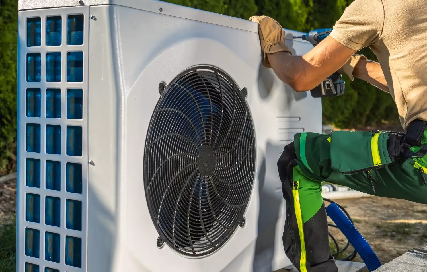 An HVAC technician in green work pants and gloves kneels beside a white outdoor heat pump unit. He uses a power drill to secure or remove the side panel of the system.
