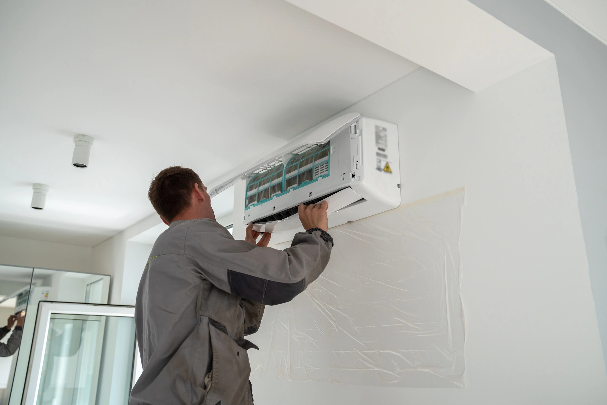 A technician in a gray uniform services an air conditioner mounted on a white wall.
