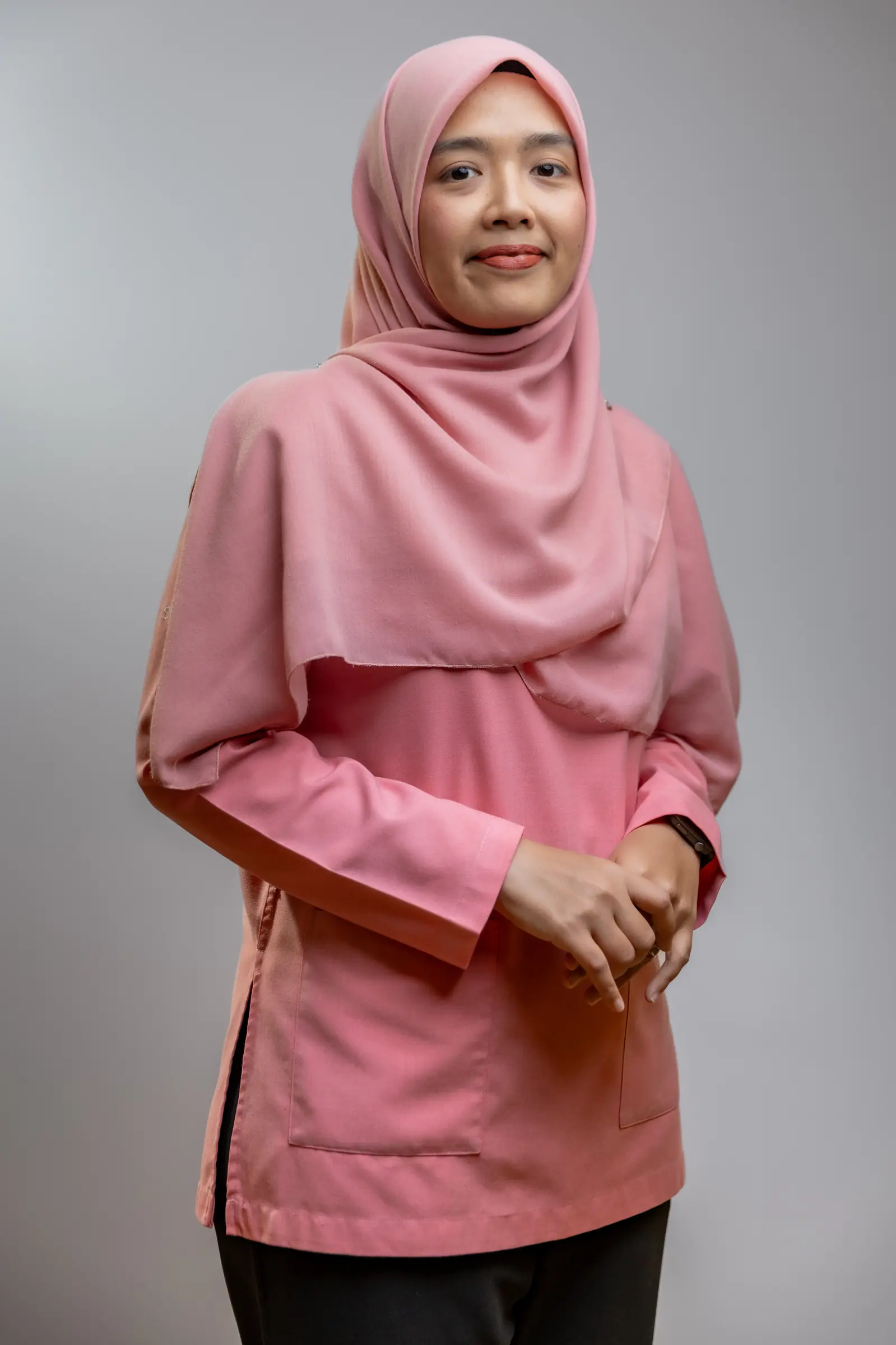Woman wearing a pink hijab and matching long-sleeve top standing against a gray background.
