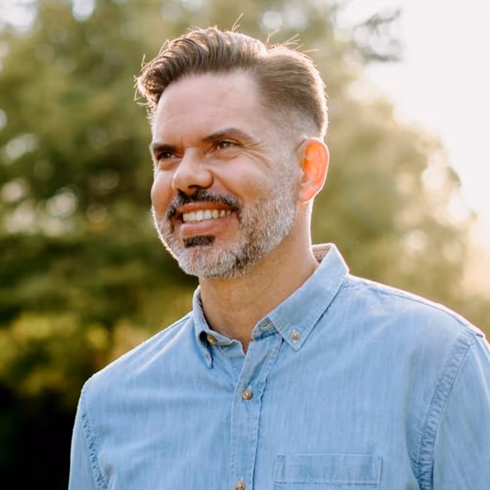 Smiling middle-aged man with short hair and salt-and-pepper beard wearing a light blue button-up shirt outdoors.