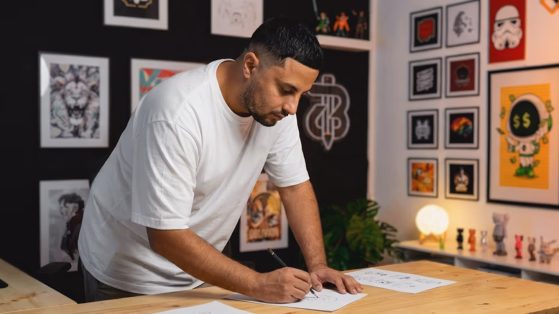 Man in a white t-shirt leaning over a wooden table, drawing on paper with a pencil in a room decorated with framed artwork.