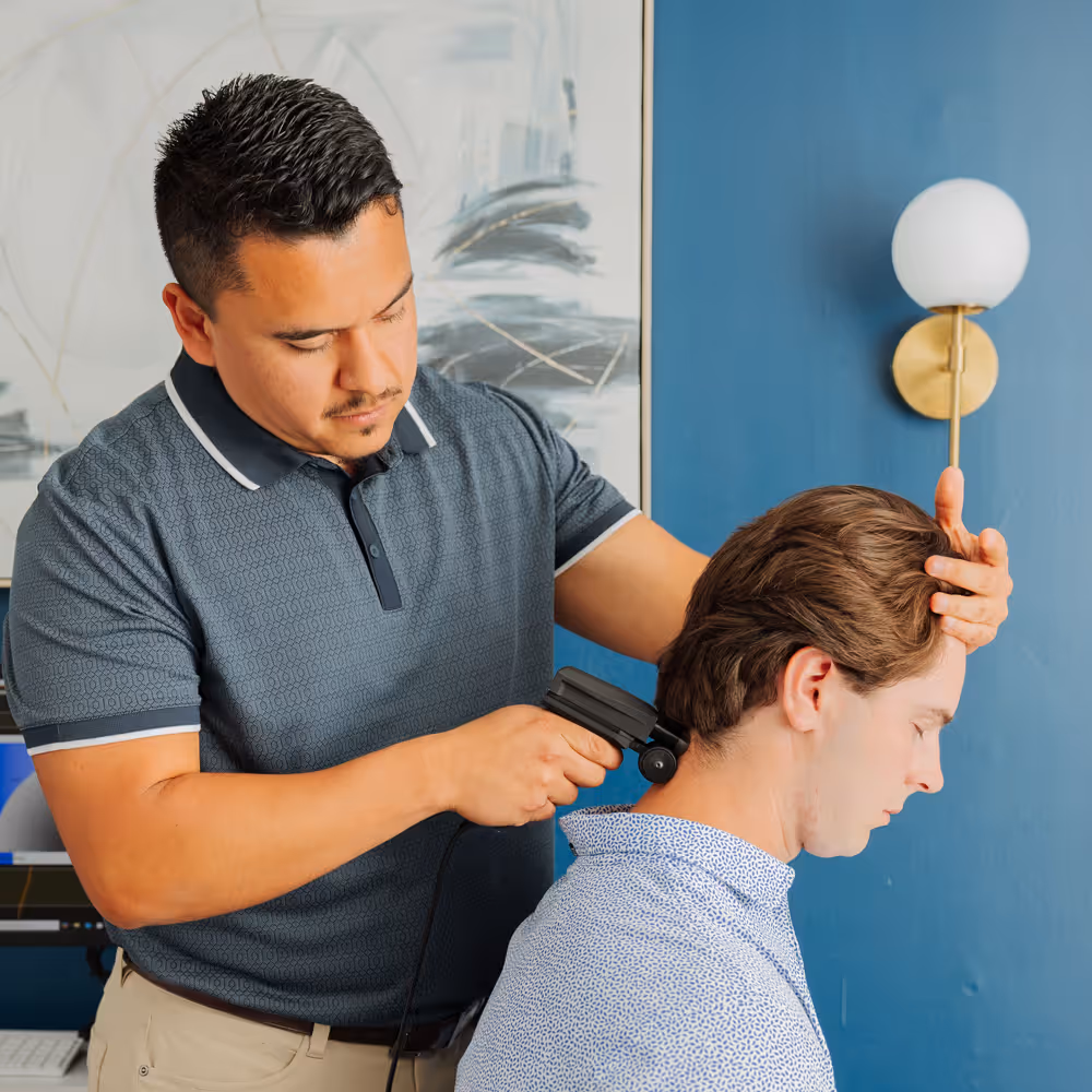 Chiropractor using a handheld percussion massager on a seated man's neck in a blue-walled office.