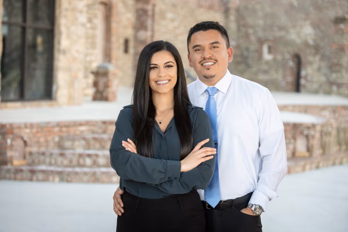 Smiling man in white shirt and blue tie standing behind a woman with long dark hair and crossed arms in front of a brick building.