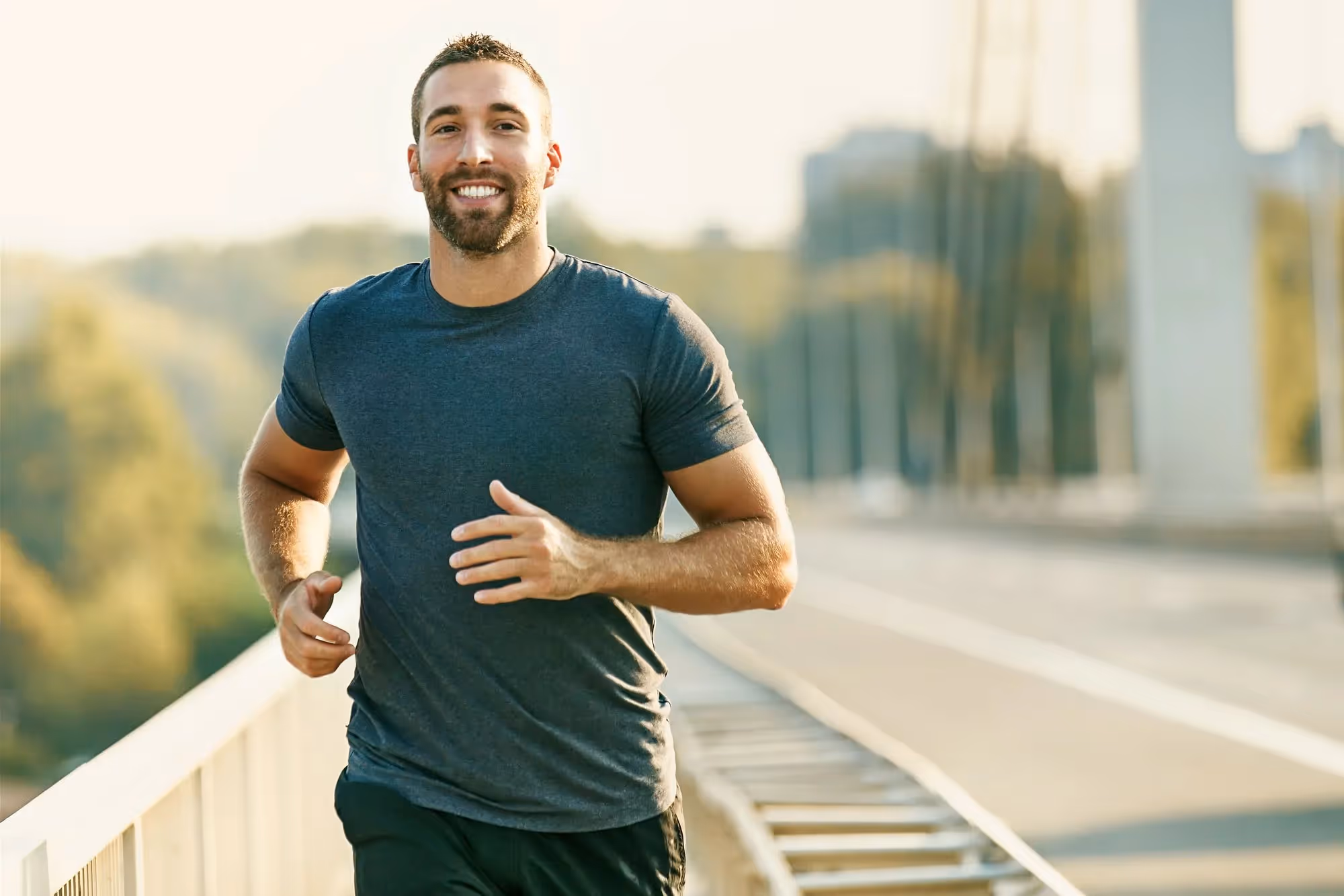 Smiling man jogging outdoors on a bridge in daylight wearing a gray t-shirt and black shorts.