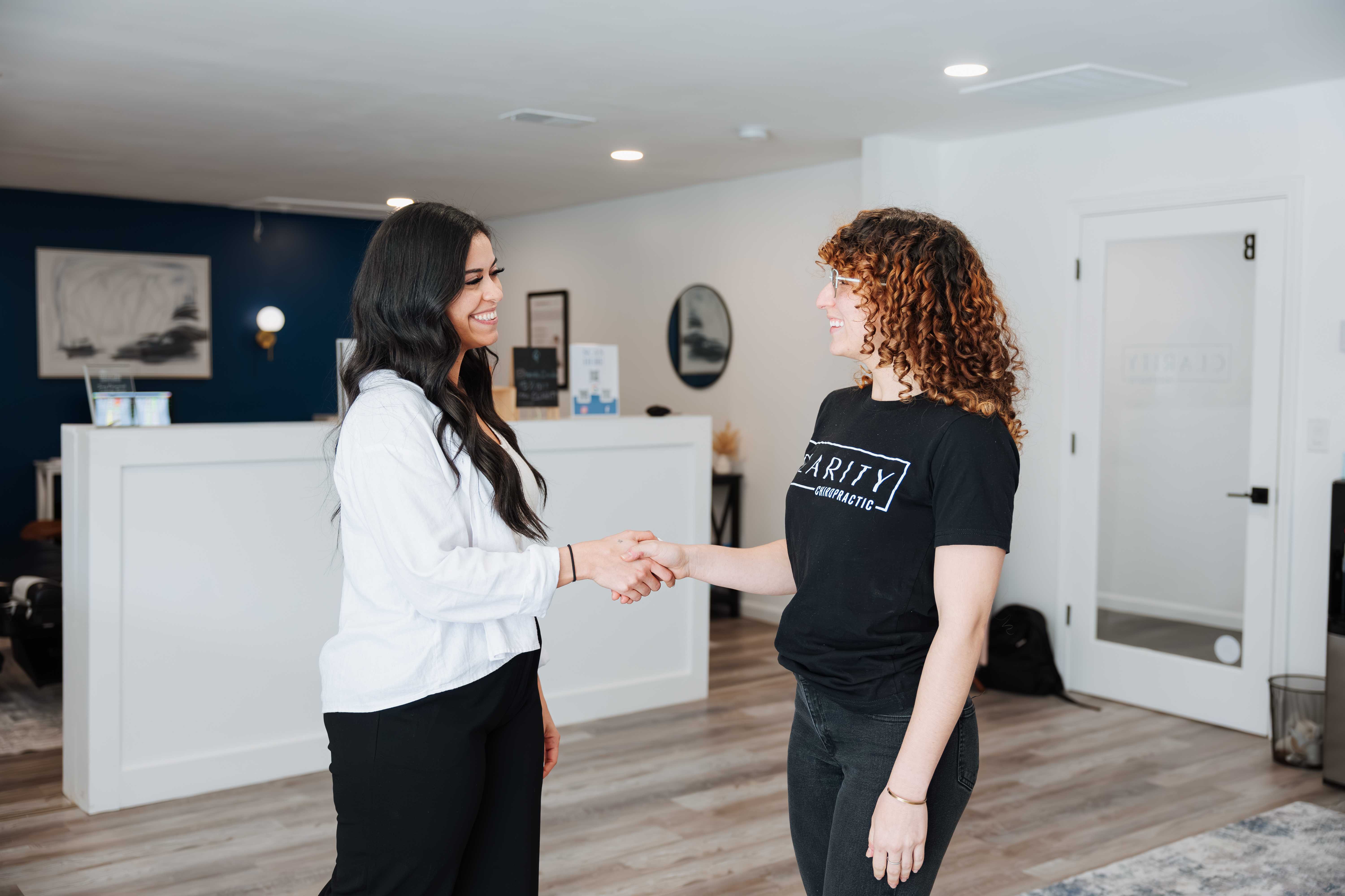 Two women smiling and shaking hands inside a modern chiropractic office reception area.