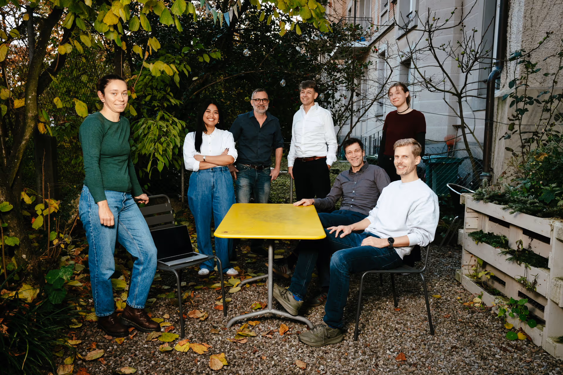 Gruppenfoto eines Teils der Belegschaft von STURM UND BRÄM. Von Links nach Rechts: Mena Lerf, Aira Manalo, Yves Thomi, Urs Bräm, Bernhard Sturm, May Kulowatz, Aram Losmann. Die Personen sitzen draussen um einen gelben Gartentisch.