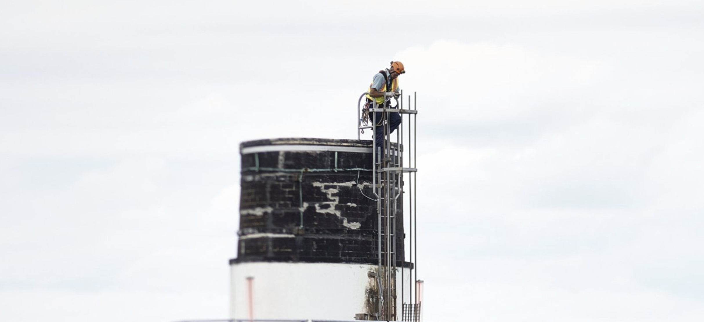 Man sieht einen Industriekamn vor grauem Himmel. Zuoberst ist ein Mann mit Helm und Kletterausrüstung zu sehen.