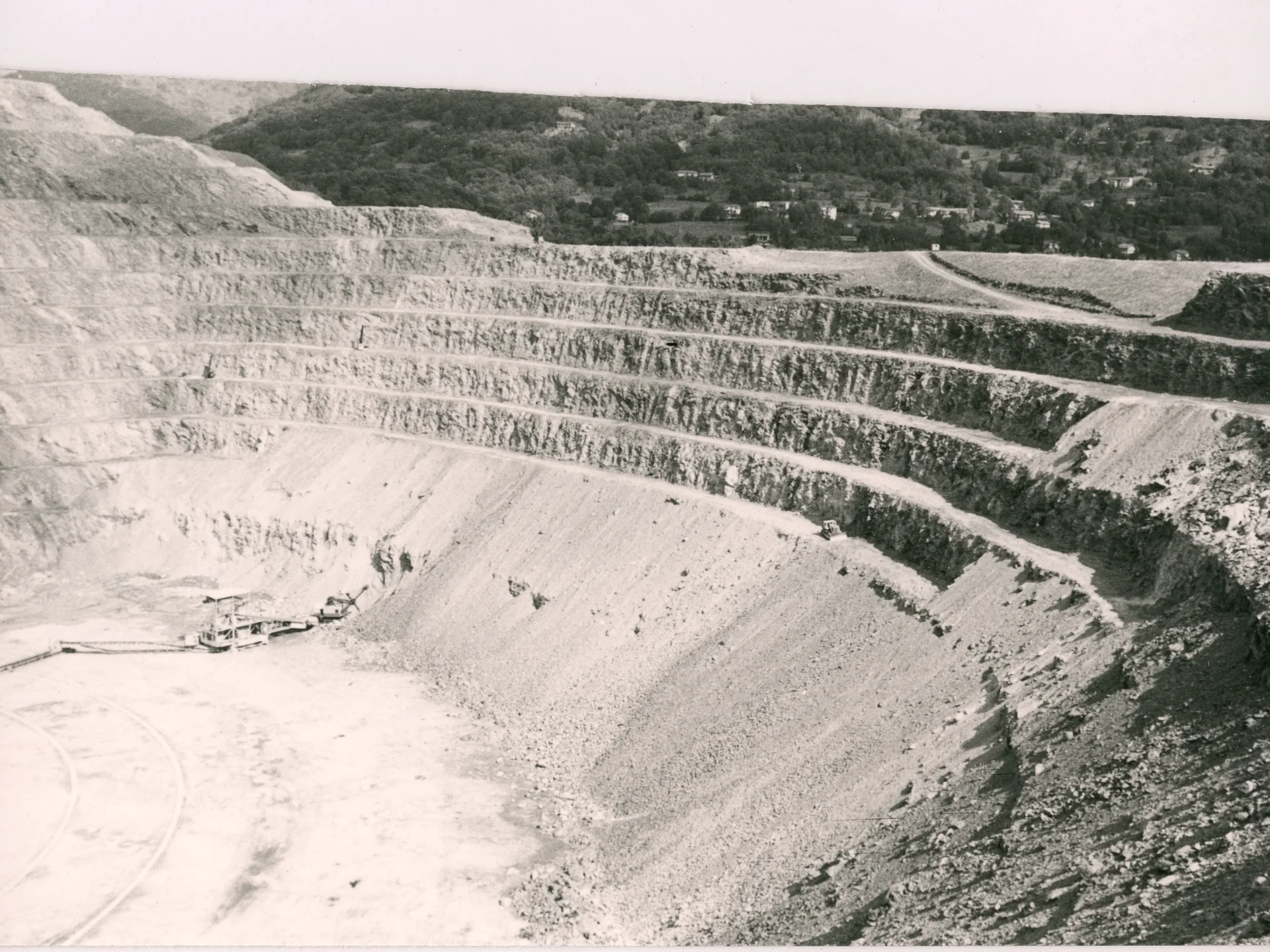 Terraced open-pit mine with machinery at the bottom and green hills in the background.
