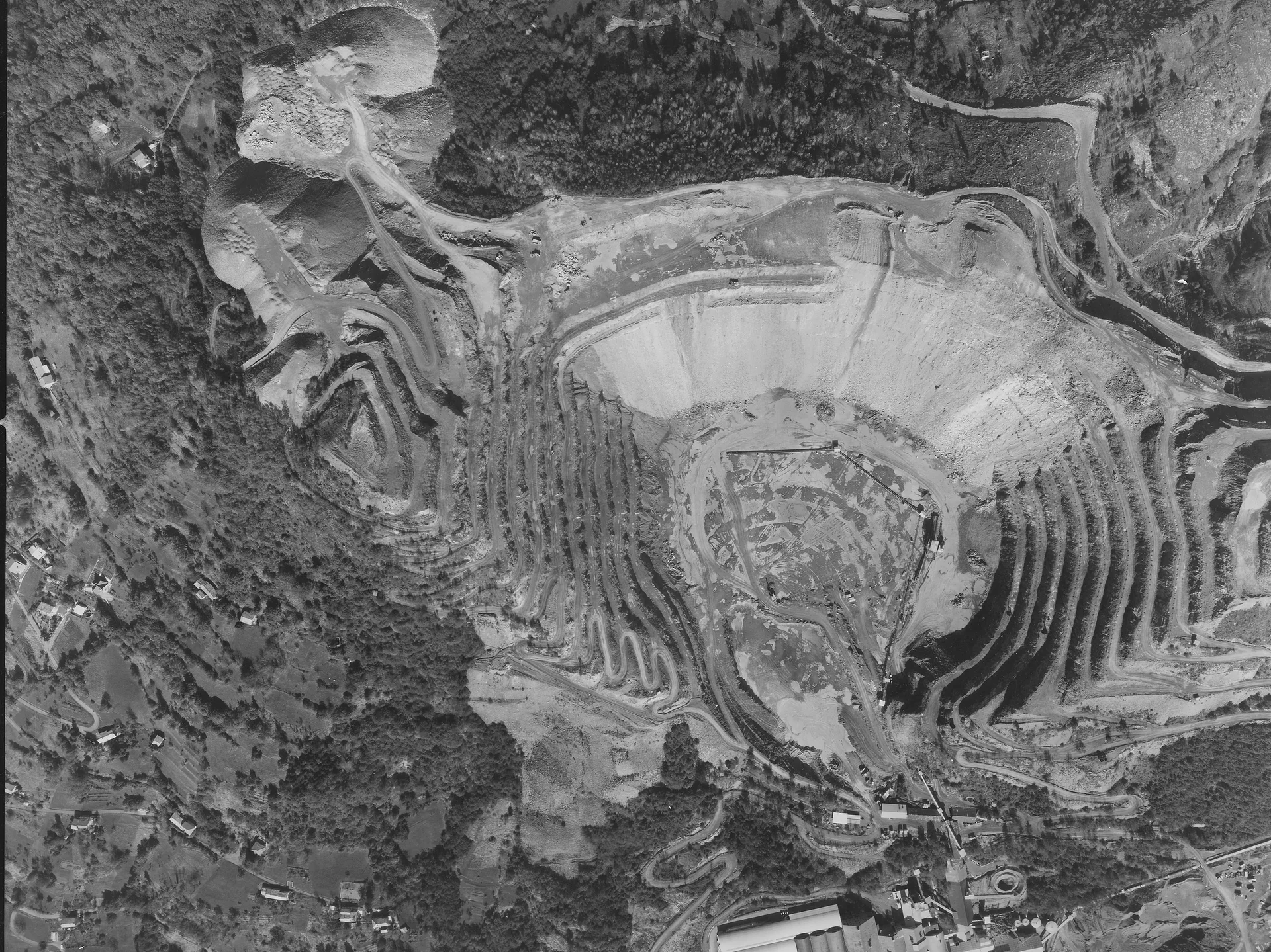 Aerial view of a large terraced open-pit mine surrounded by forest and scattered buildings.