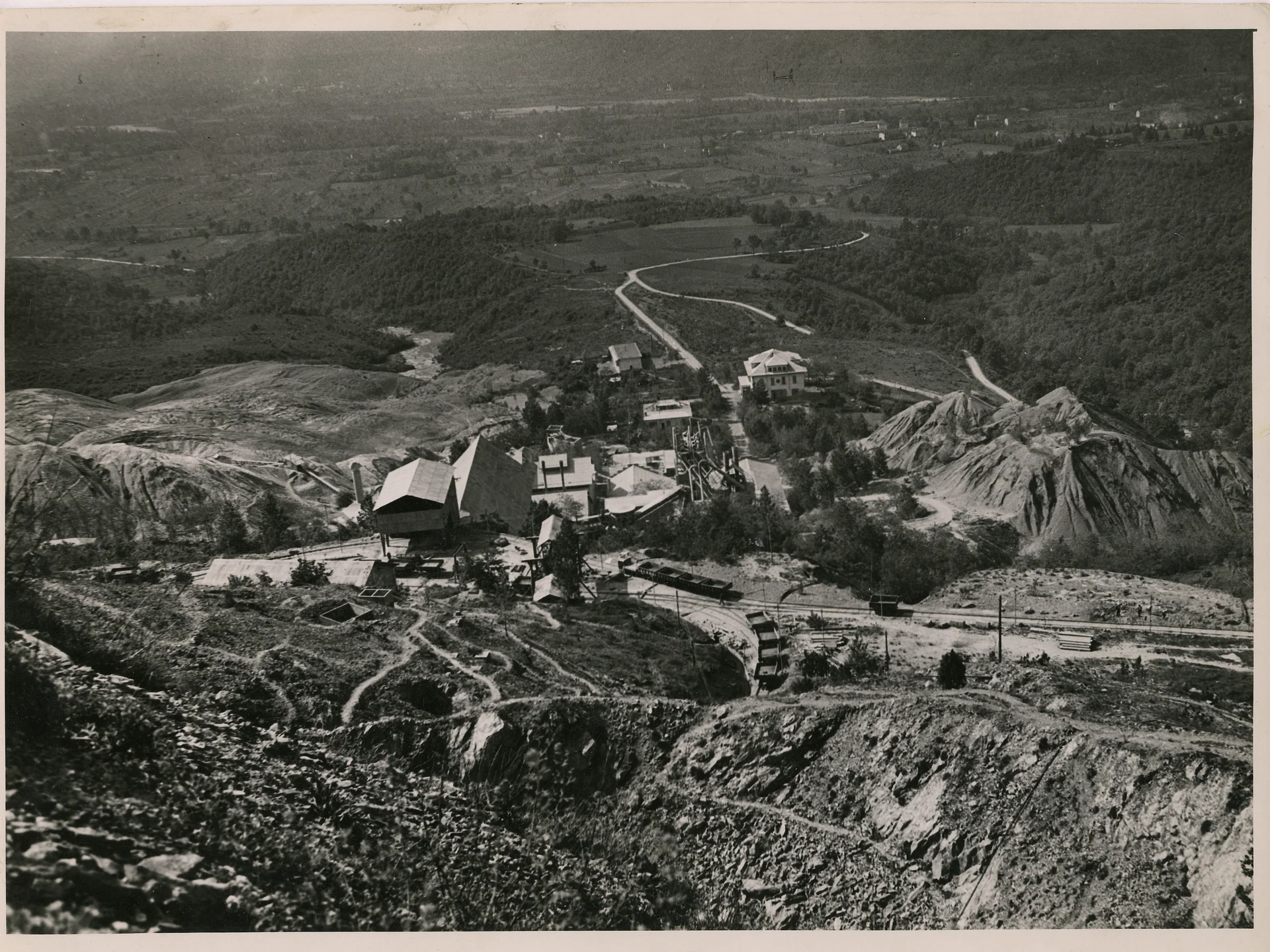Black and white aerial view of a mining site with buildings, equipment, and rail tracks in a rural landscape.