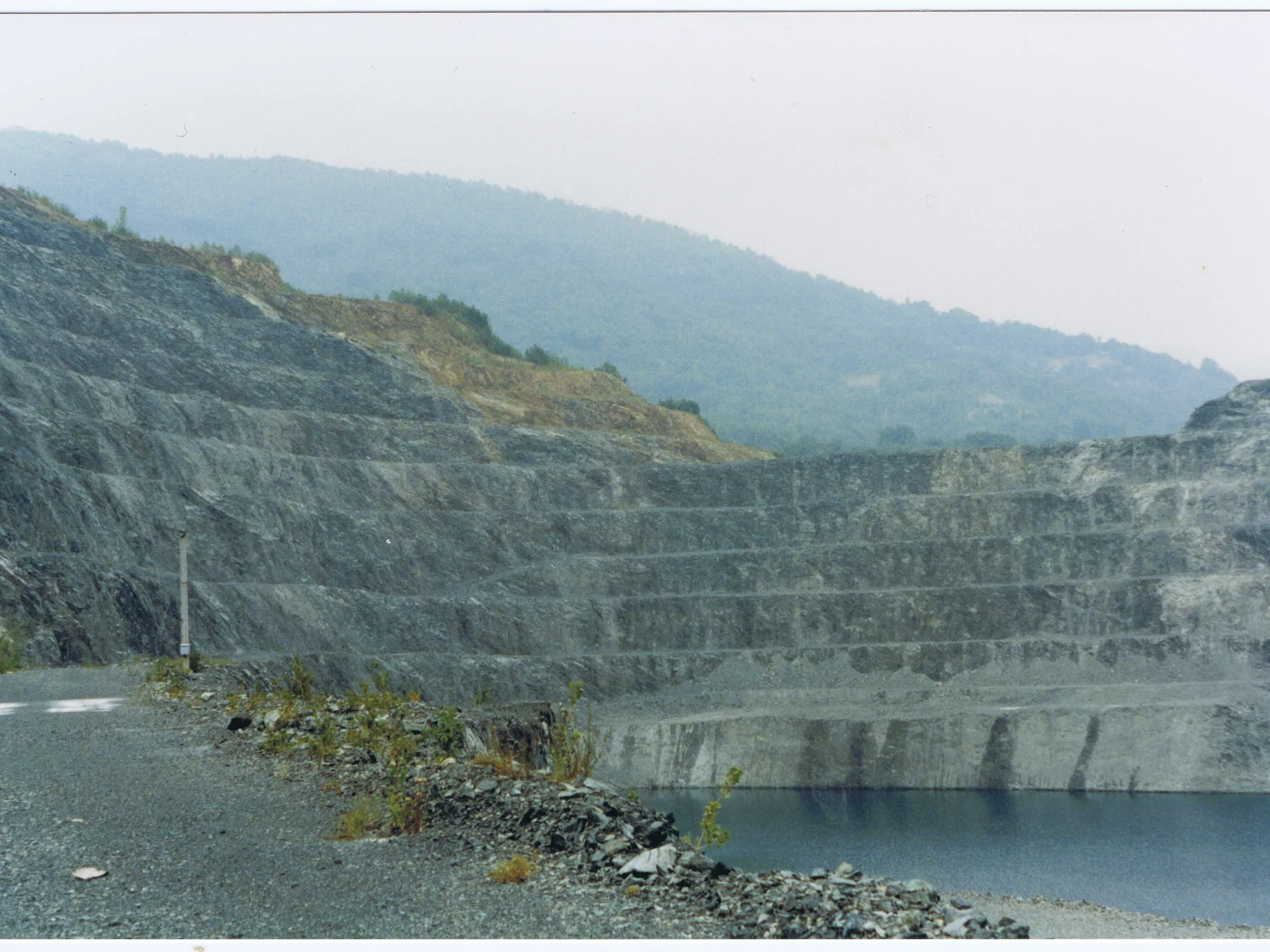 Stepped rock quarry beside a water-filled pit with hazy hills in the background.