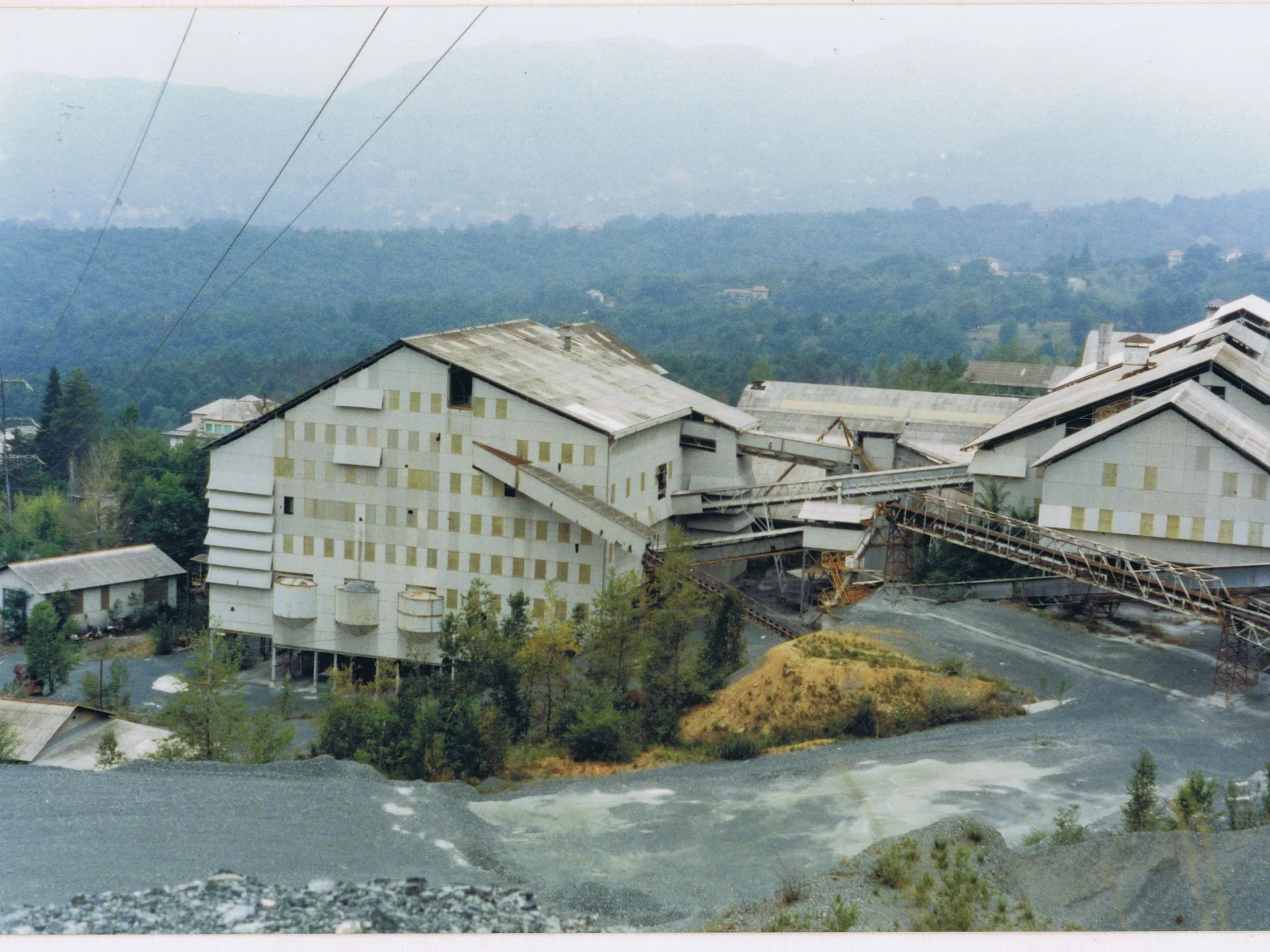 Industrial buildings with conveyors surrounded by forest and hills under an overcast sky.