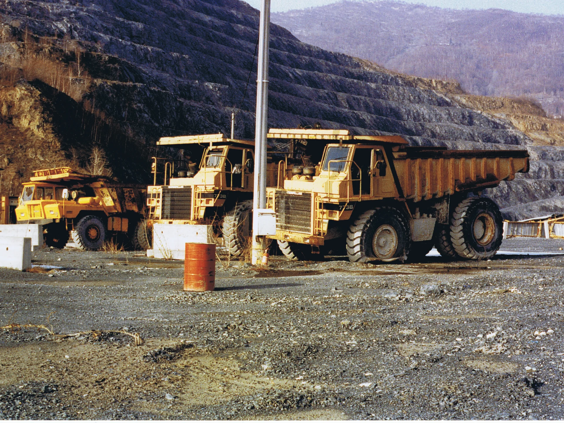 Two large yellow mining dump trucks parked on gravel with terraced hills in the background.
