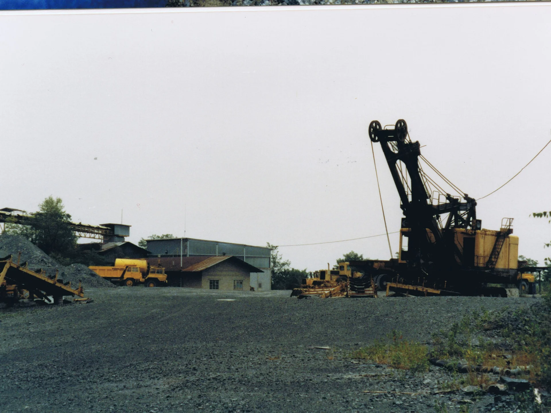 Mining site with large black and yellow excavator, yellow trucks, small buildings, and gravel ground.