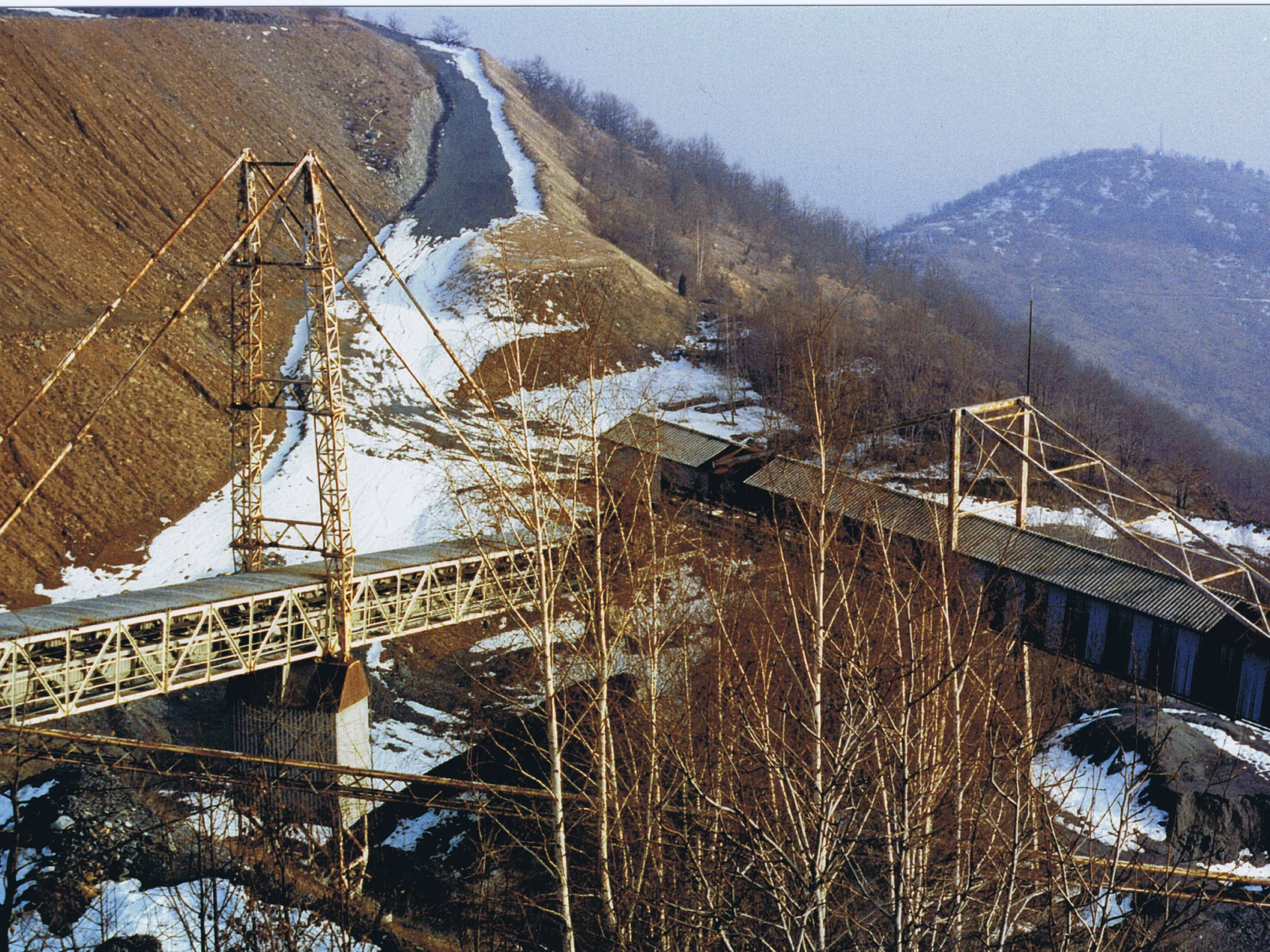 Two metal truss bridges crossing a snowy valley with barren trees and hills in the background.