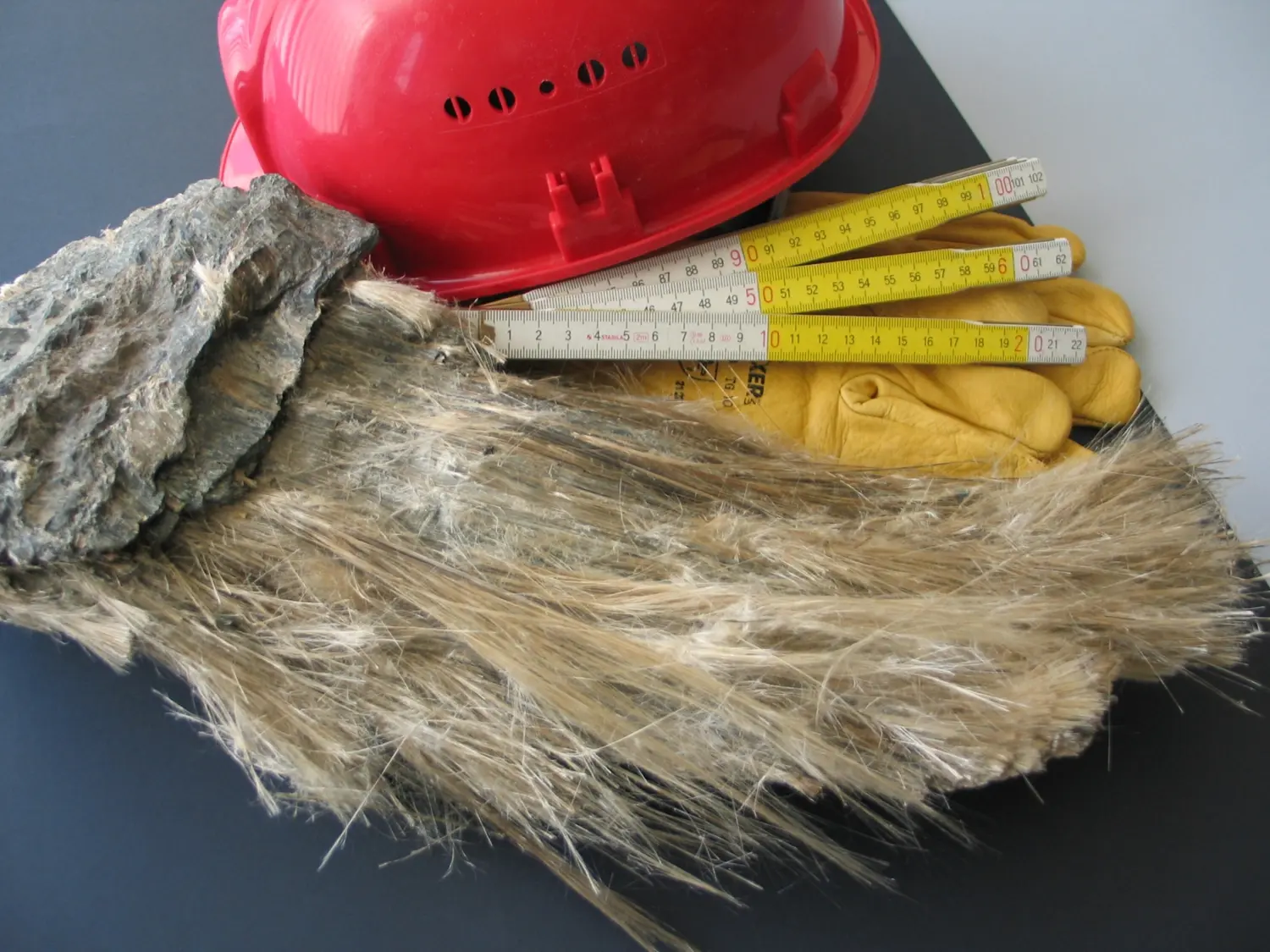 Campione di amianto fibroso accanto a casco di sicurezza, guanti da lavoro e metro pieghevole su piano scuro