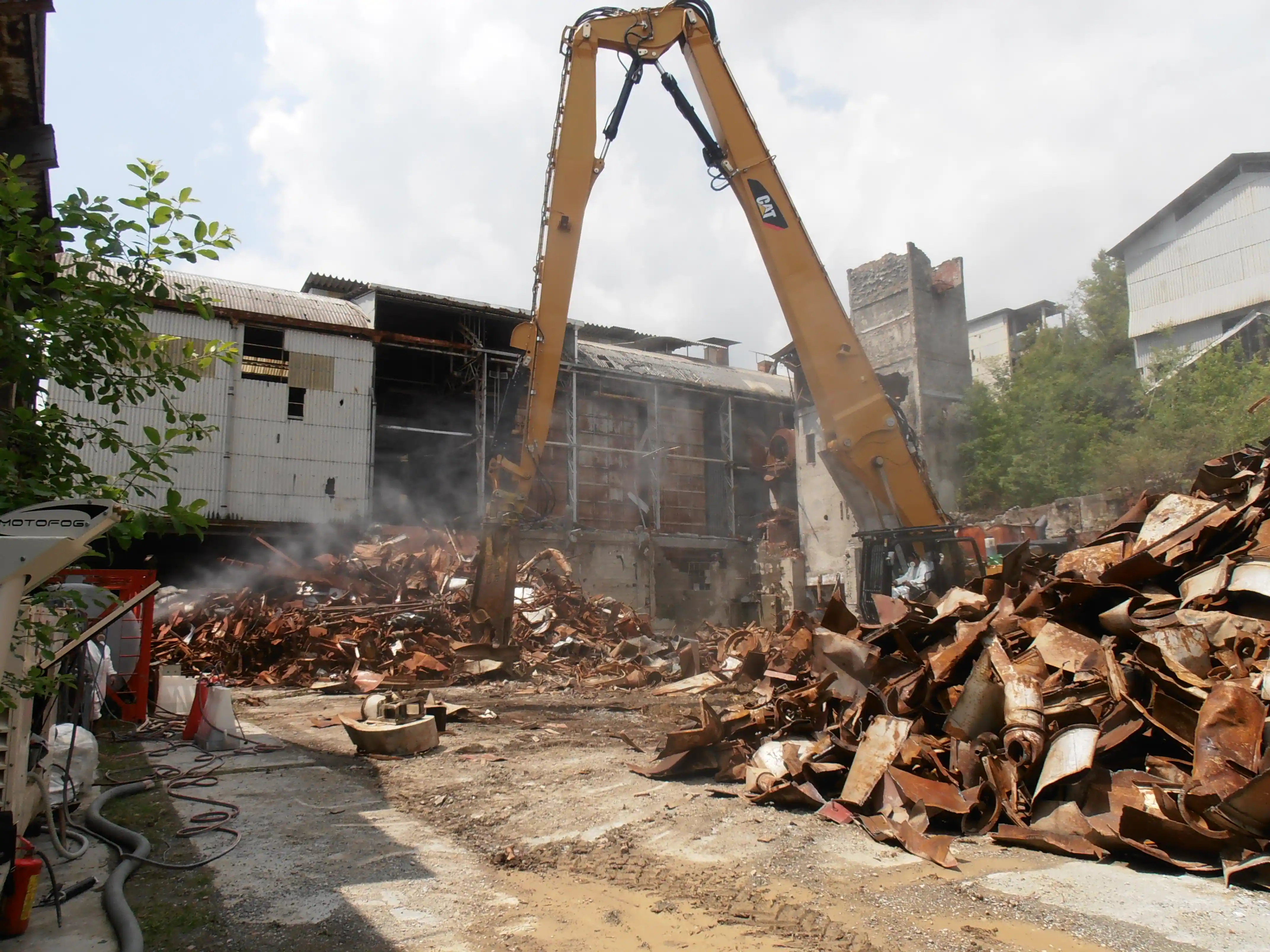 Pinza idraulica gialla che taglia rottami metallici e travi arrugginite in un cantiere di demolizione