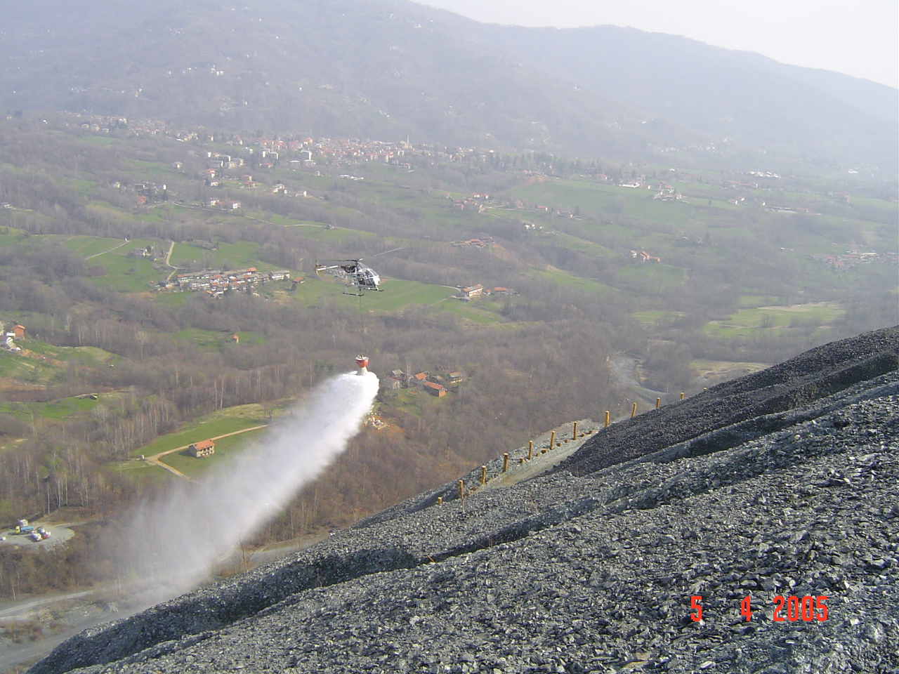 Elicottero in volo distribuisce miscela acquosa con sementi e concimi su un ripido pendio di detriti grigi sopra una vallata con centri abitati