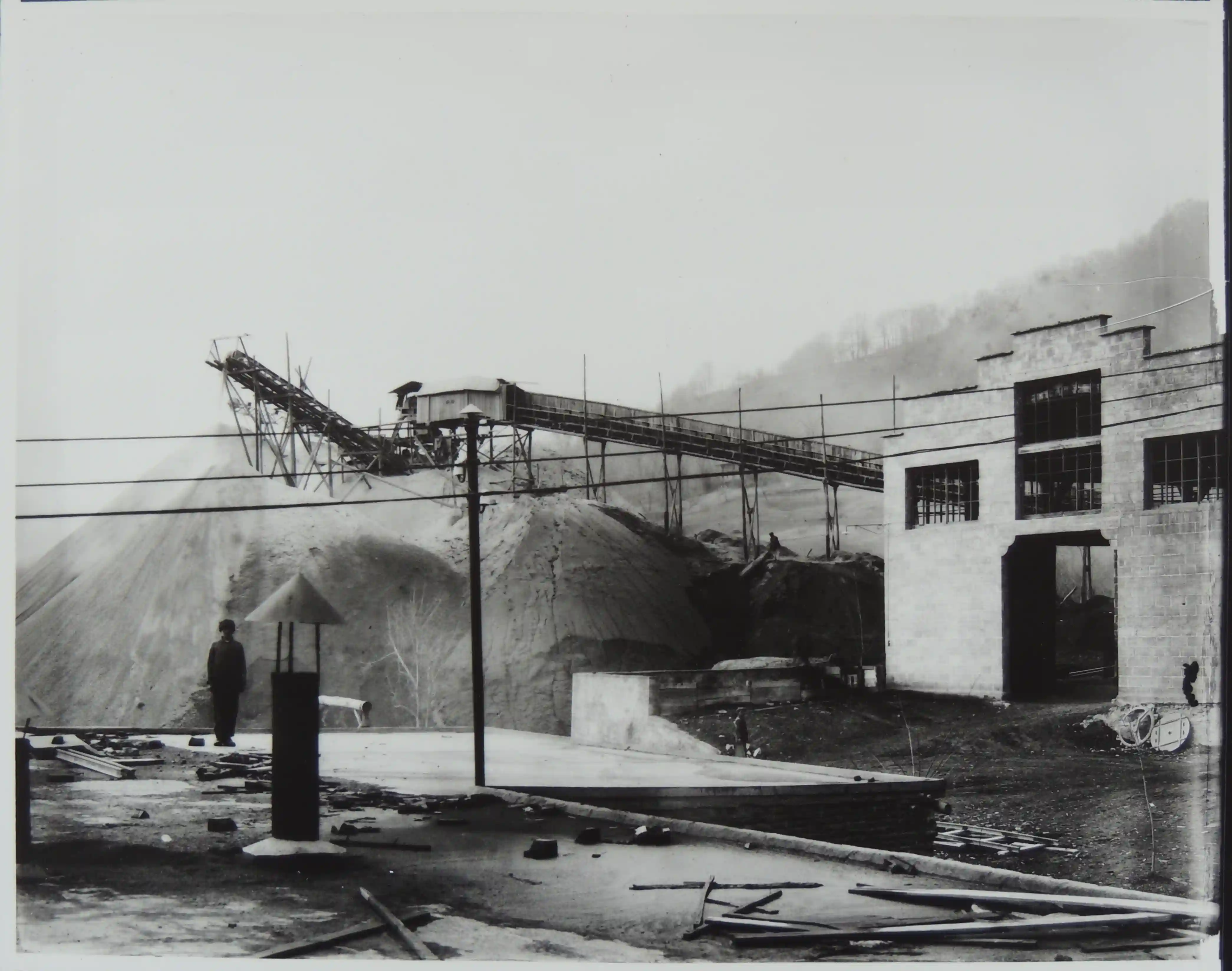 Nastro trasportatore d'epoca sospeso sopra grandi cumuli di pietrisco scarto di produzione, accanto a un edificio con aperture simmetriche