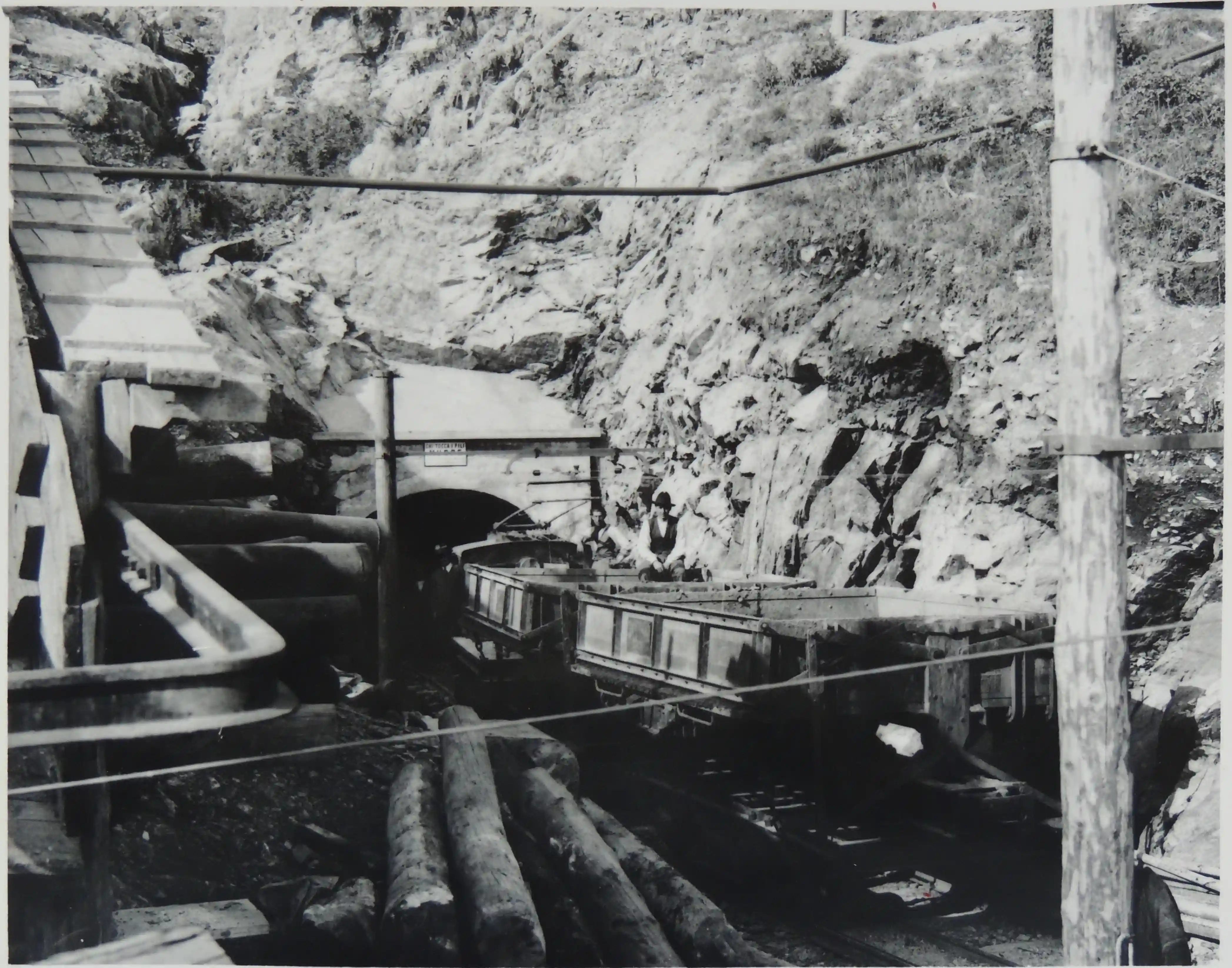  Ingresso di una galleria mineraria in roccia con carrelli su binari e tronchi di legno accatastati all'esterno