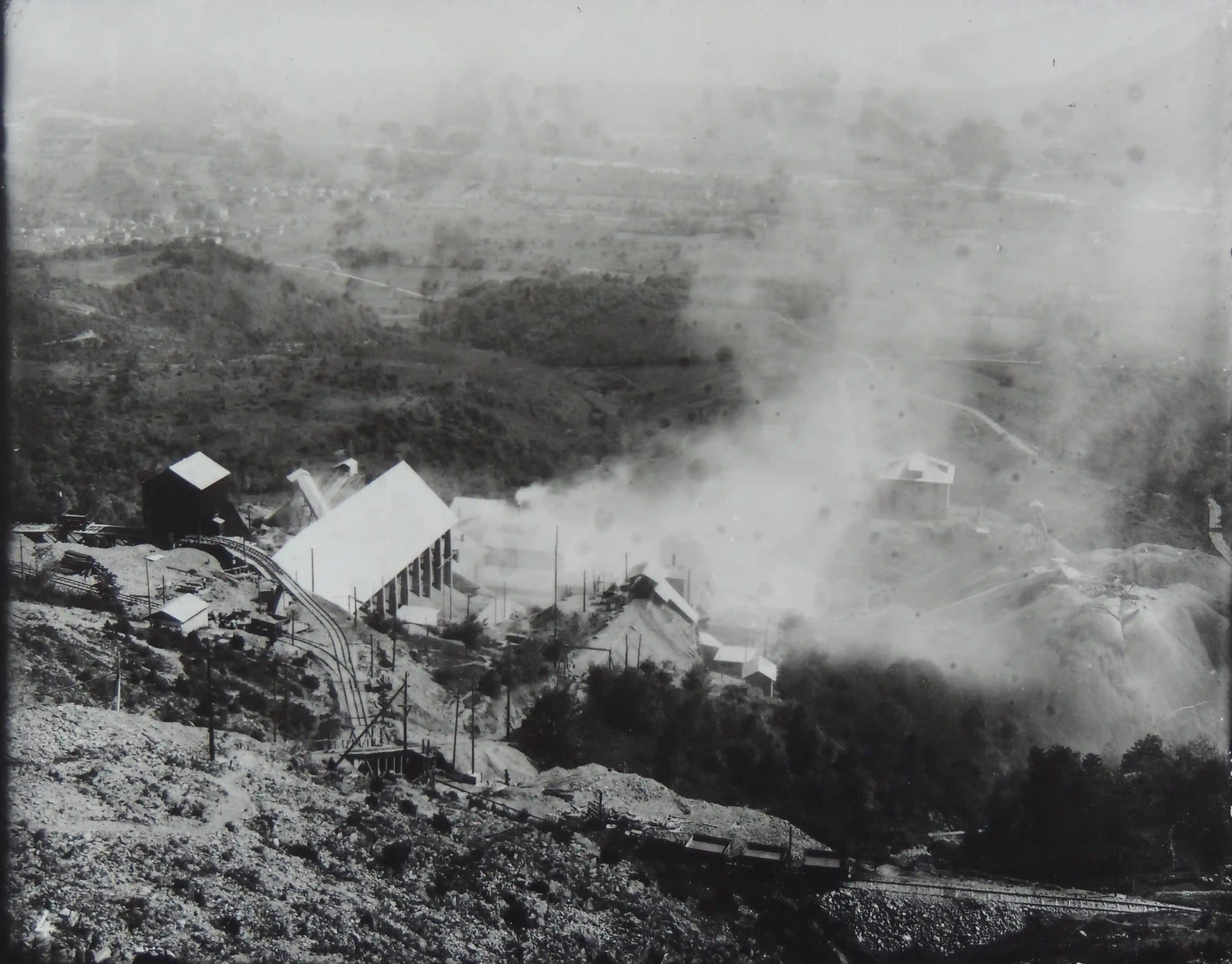 Panoramica storica del sito minerario, con gli stabilimenti a valle, caratterizzati da densi fumi industriali, edifici produttivi e binari per il trasporto dei carrelli.
