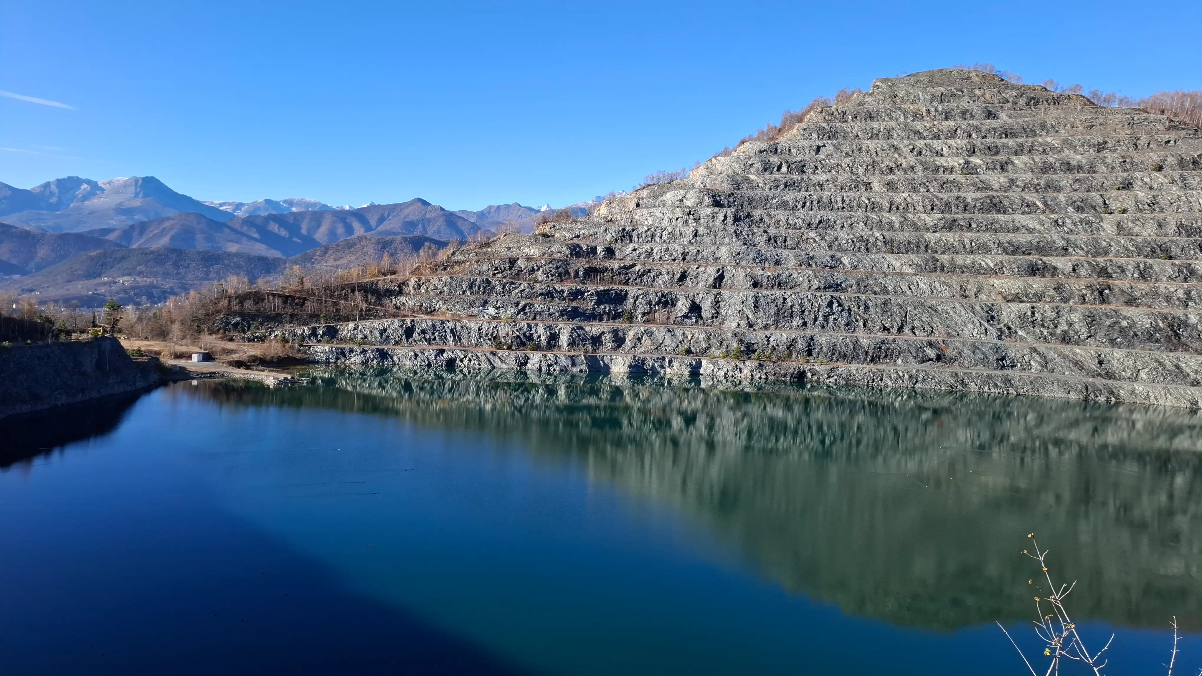 Lago ai piedi di cava terrazzata, con montagne alpine sullo sfondo e cielo limpido