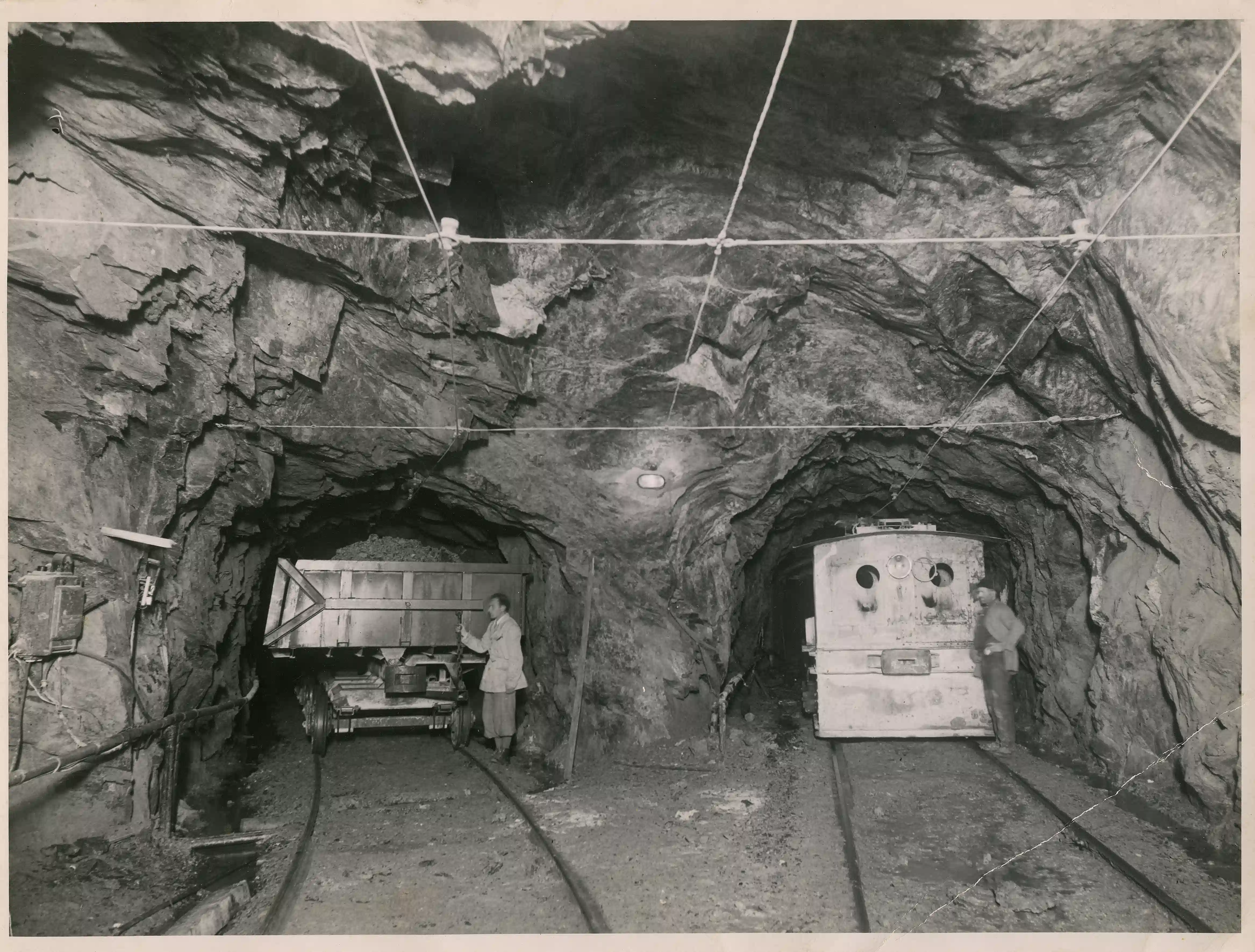 Interno di una galleria mineraria con binari, vagoncini carichi e minatori al lavoro sotto cavi elettrici sospesi