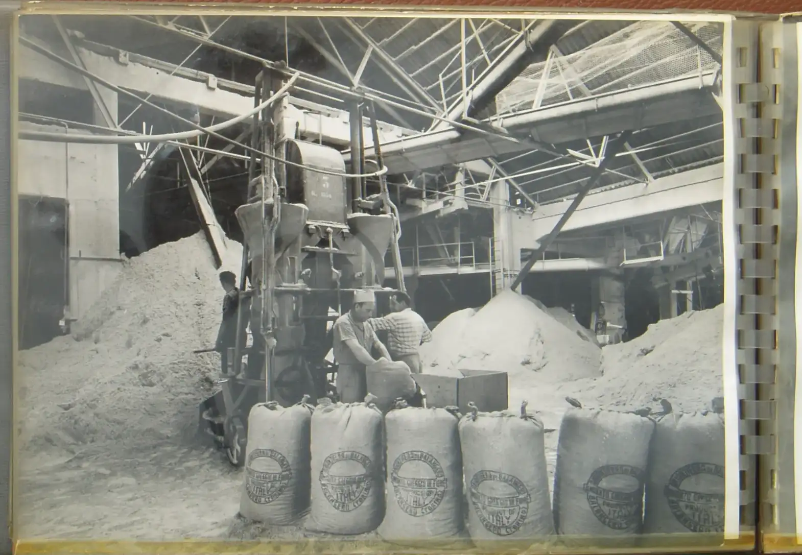 Interno di fabbrica d'epoca con operai che caricano a mano sacchi di amianto greggio davanti a grandi cumuli di minerale