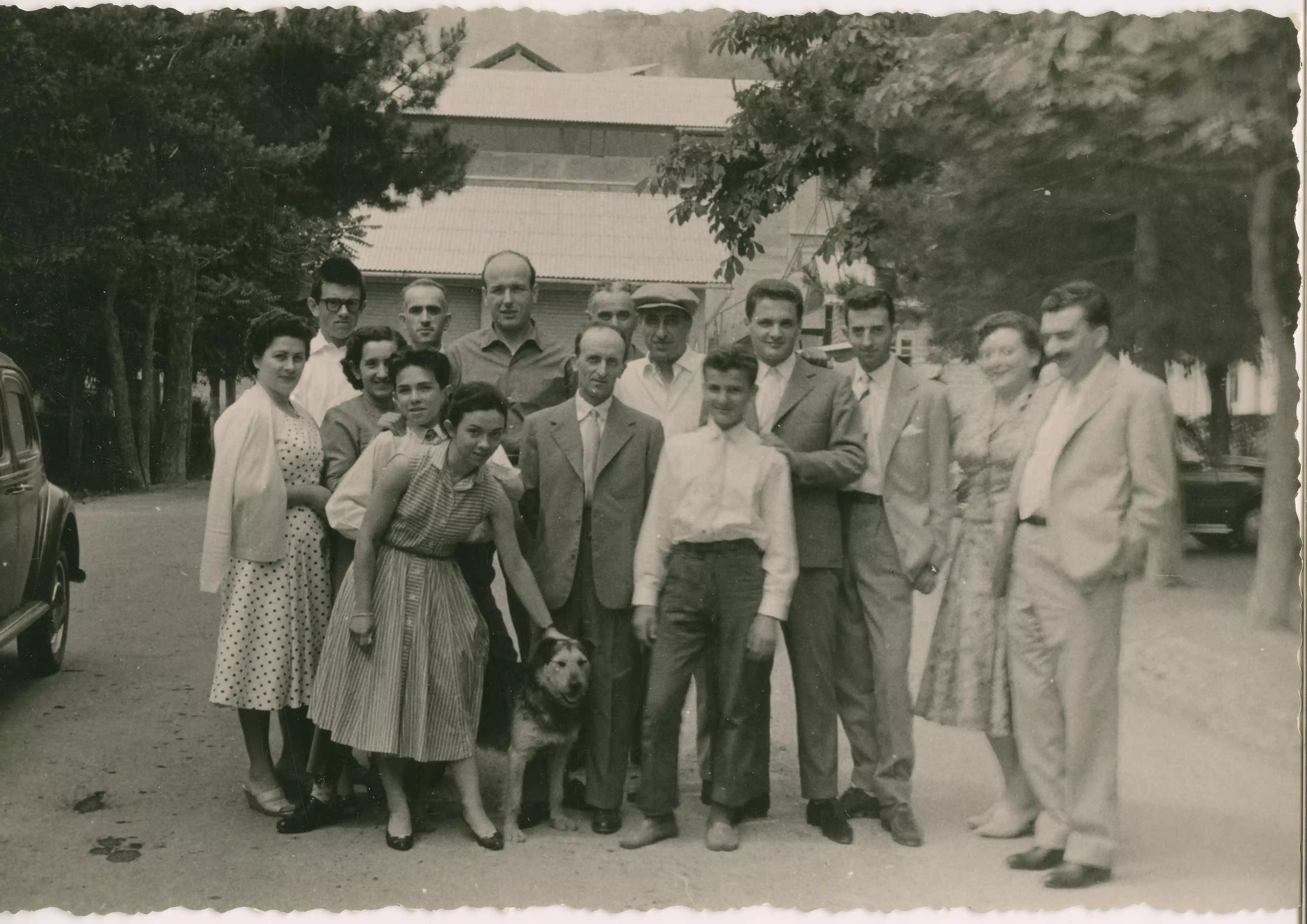 Gruppo di persone  in posa per lo scatto fotografico con un cane, databile anni  60 con sullo sfondo i capannini dello stabilimento 