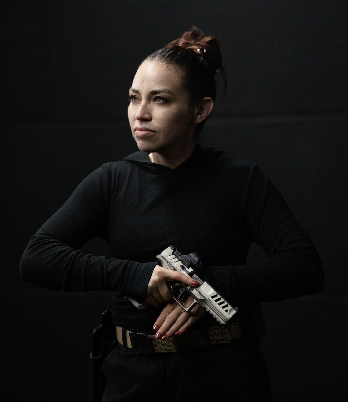Woman in black outfit holding a white handgun with both hands against a dark background.