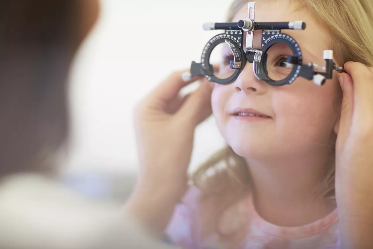Young girl smiling during an eye test, wearing a trial frame while an optometrist checks her vision