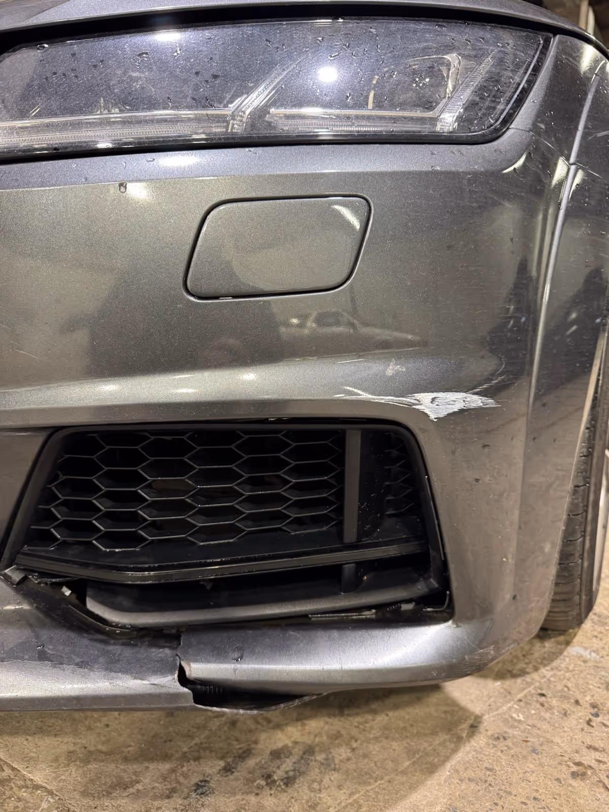 Front right corner of a gray car with a damaged bumper showing a crack, scrape, and broken grille.