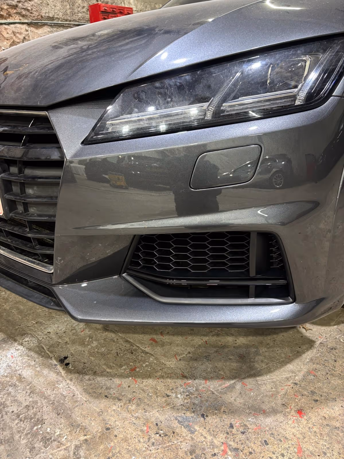 Close-up of a gray car's front left side showing the headlight, grille, and bumper in a garage.