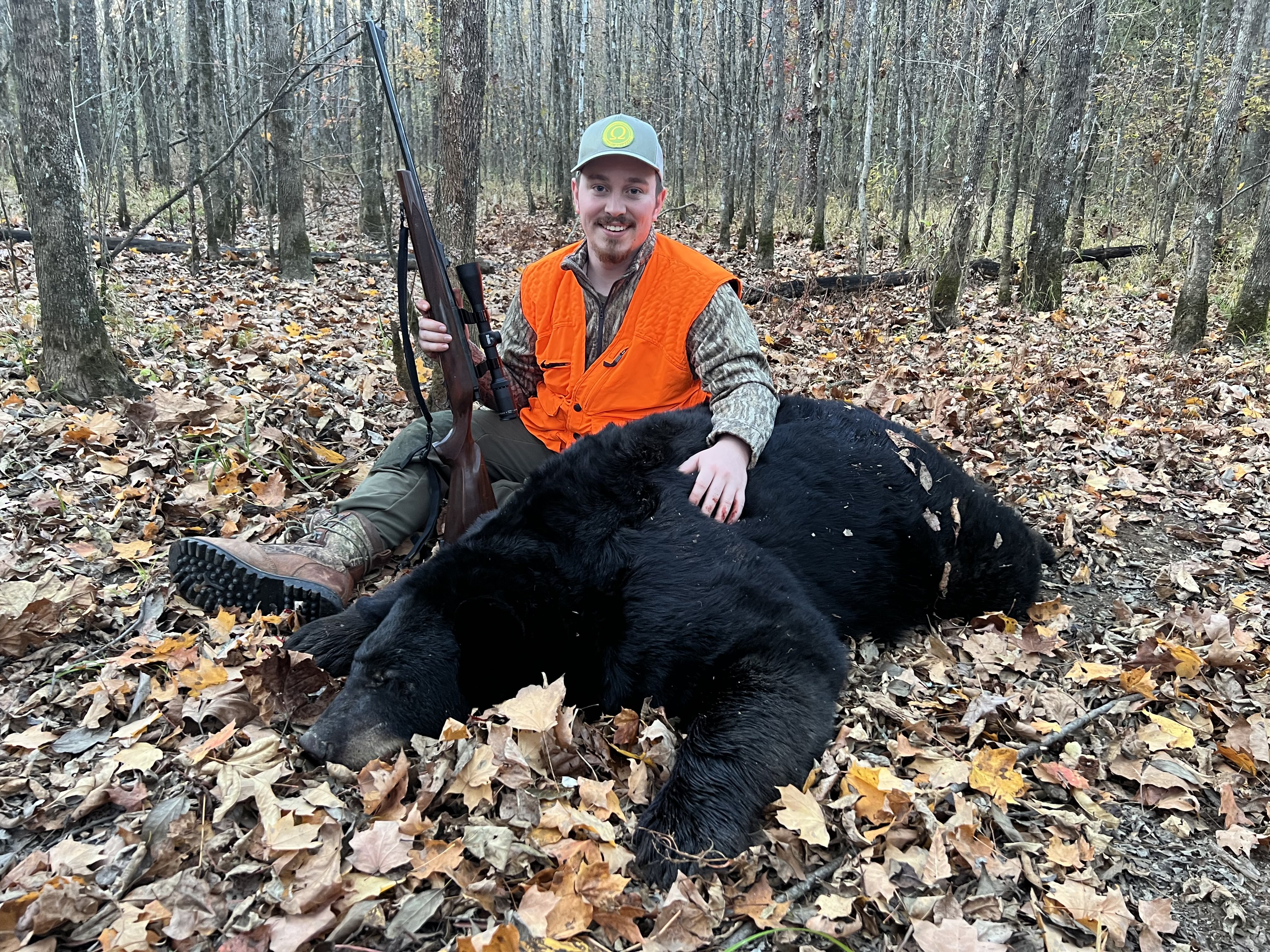 Hunter in orange vest sitting with a rifle next to a large black bear lying on autumn leaves.