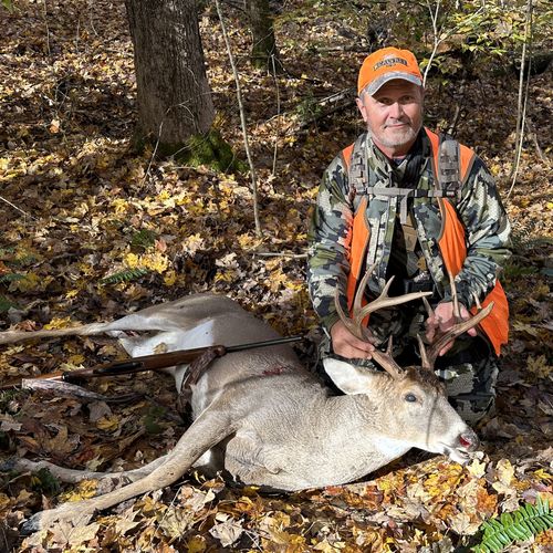 Hunter in camouflage and orange vest kneels behind a white-tailed buck with a rifle on fallen leaves.