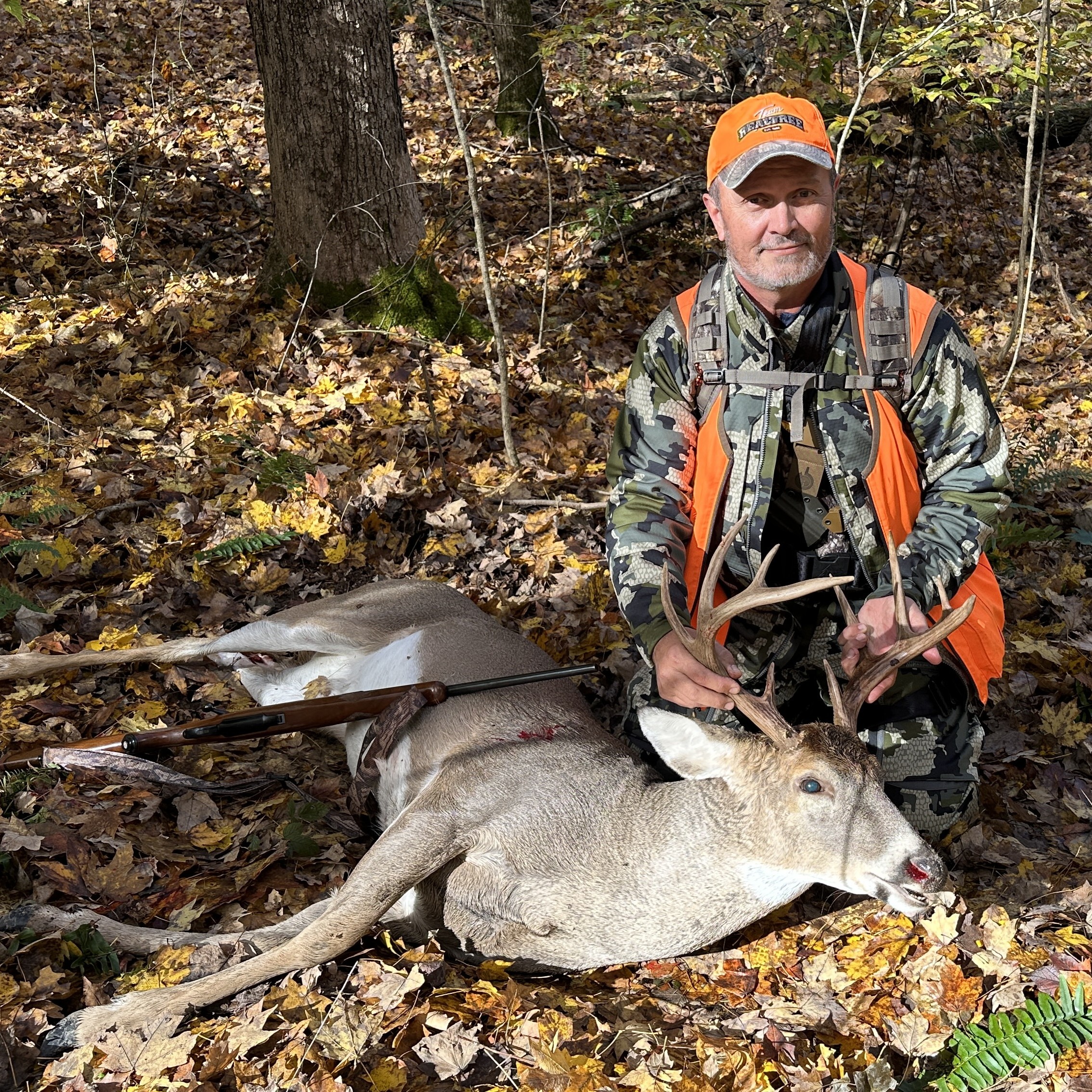Hunter in camouflage and orange vest kneels behind a white-tailed buck with a rifle on fallen leaves.