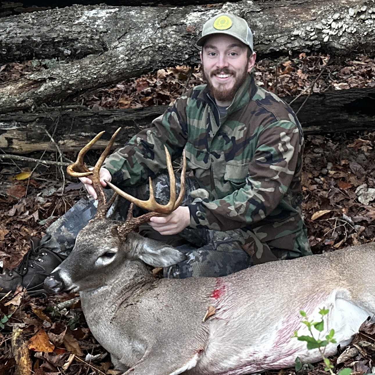 Man in camouflage holding antlers of a deer lying on the forest ground with fallen leaves.