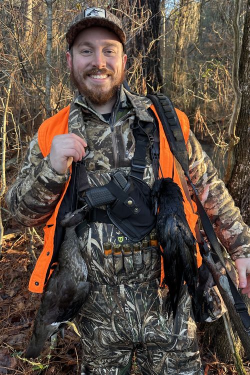 Smiling hunter in camo and orange vest holding two birds in a forest with hunting gear and shotgun.