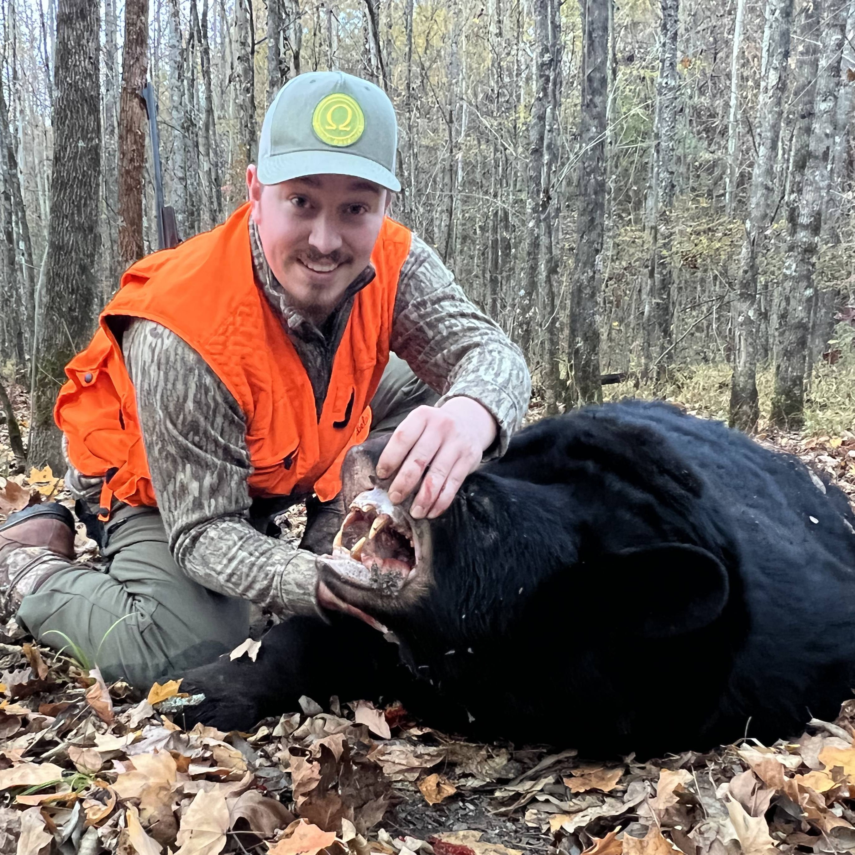 Smiling hunter in camouflage and orange vest posing with a large black bear in a forest.