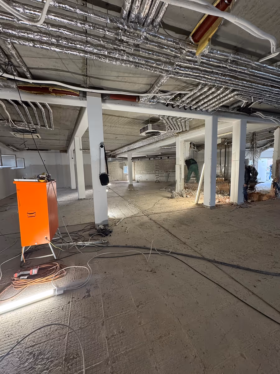 Innenraum einer Baustelle mit freiliegenden Leitungen an der Decke, zwei Bauarbeitern und orangefarbenem Baustellengerät.