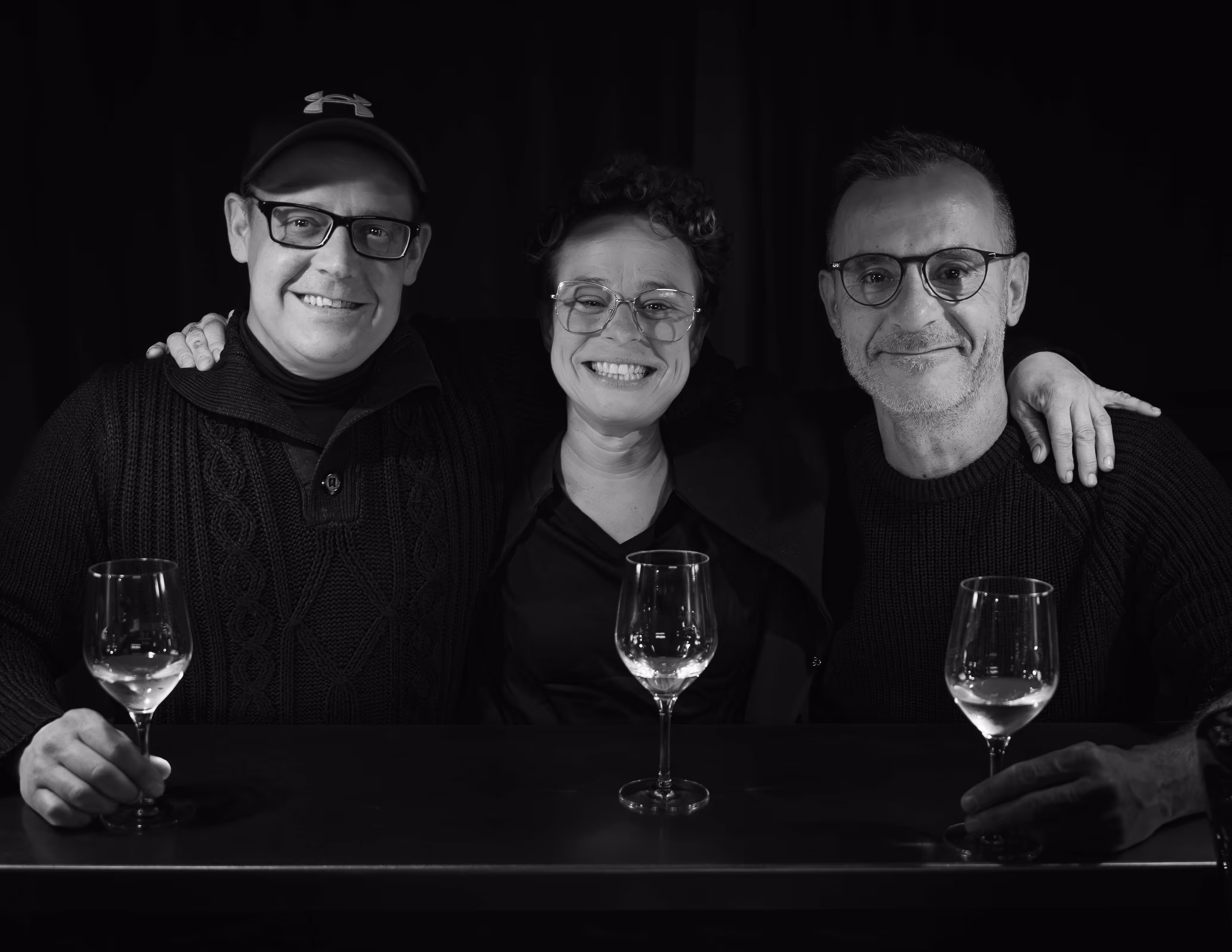 Das Barbar Team sitzt nebeneinander an der Bartheke mit Weingläsern vor sich, schwarze Hintergrund.