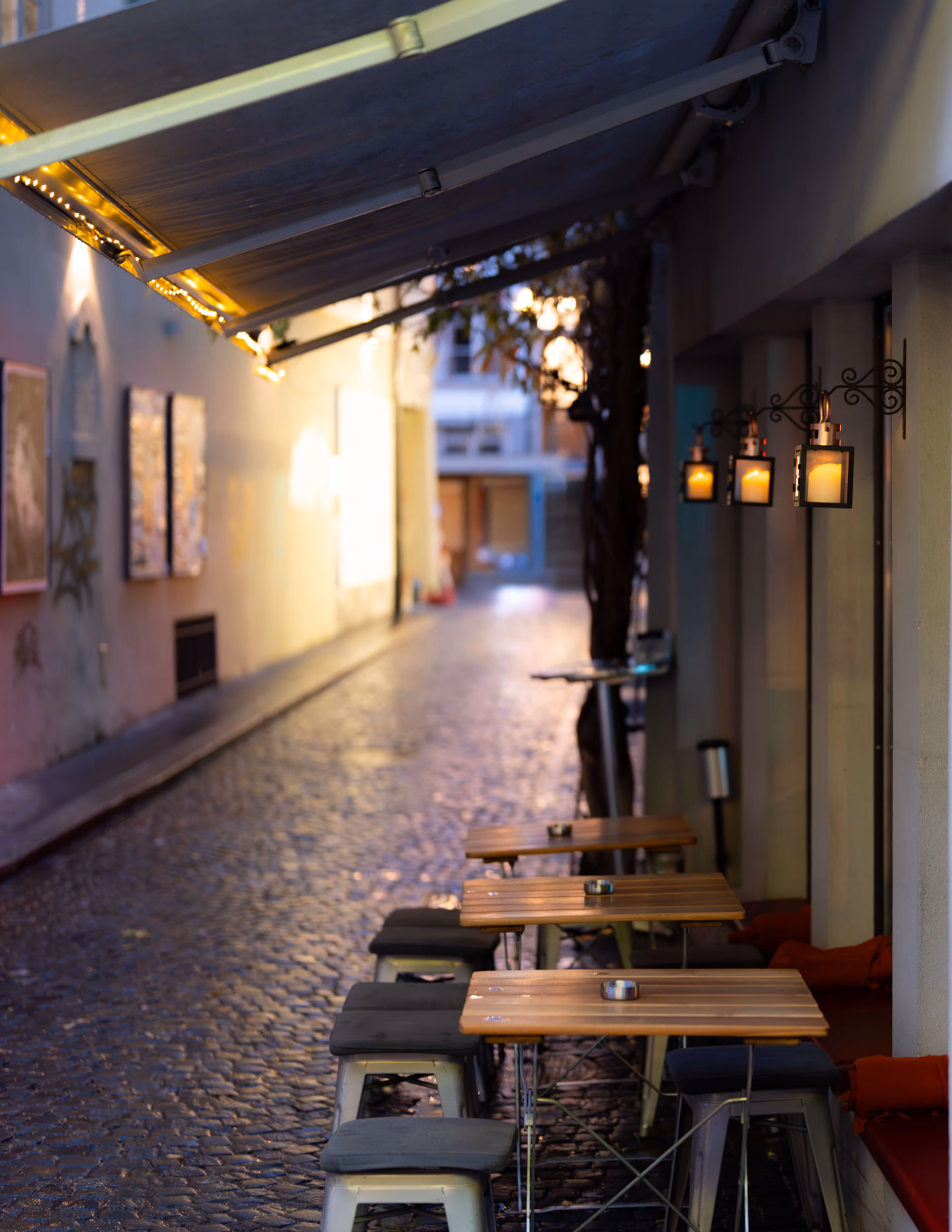 Gemütlicher Sitzbereich der Barbar mit Holzplatten und Hockern in einem gepflasterten engen Gasse bei Dämmerung.