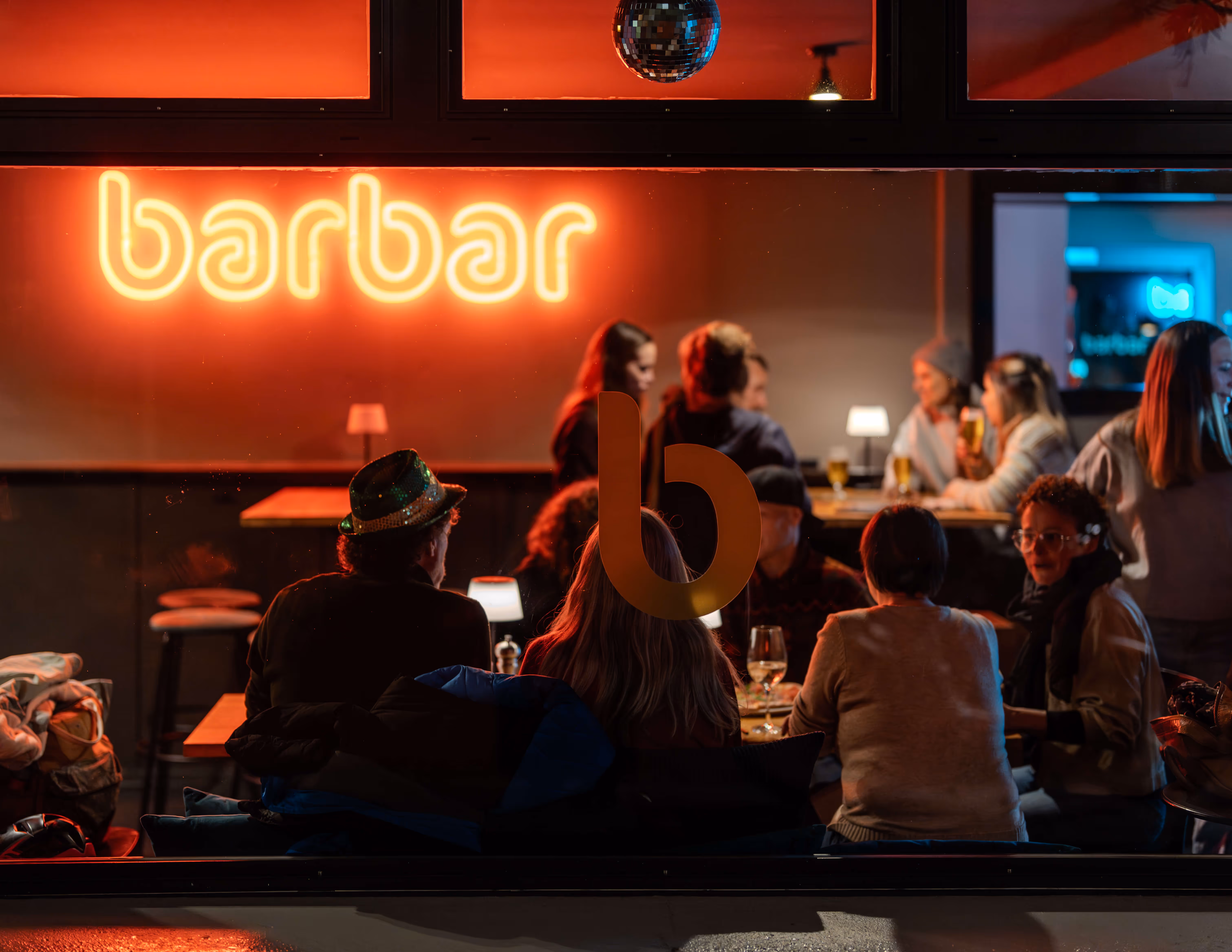 Menschen sitzen in der Barbar mit leuchtendem 'barbar'-Schild und einer Disco-Kugel an der Decke.