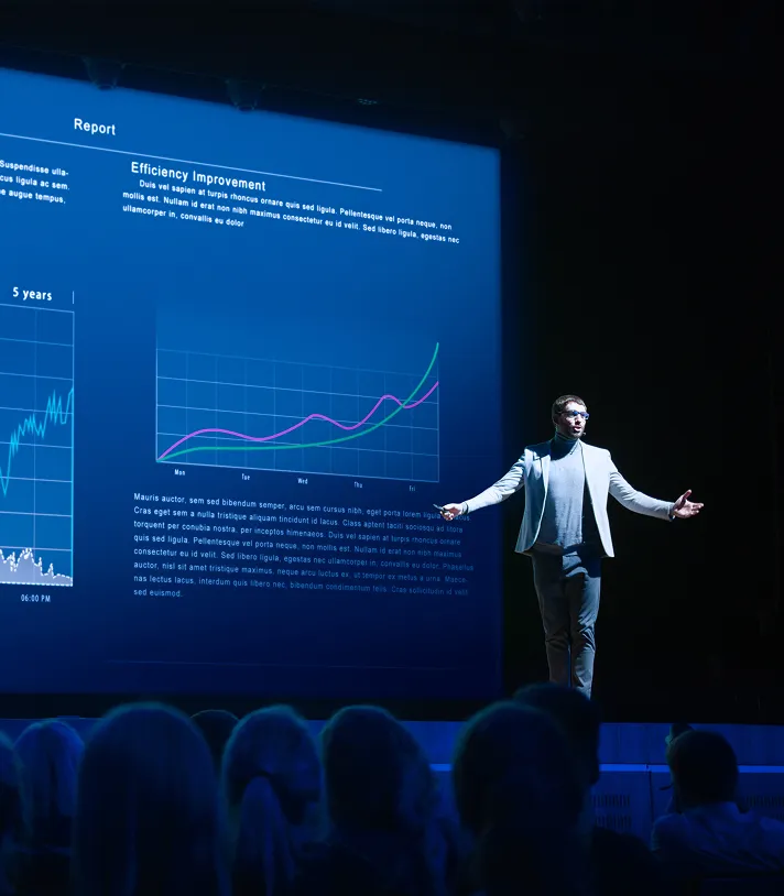 Presenter in a suit explaining efficiency improvement charts on a large screen to an audience in a dark conference room.