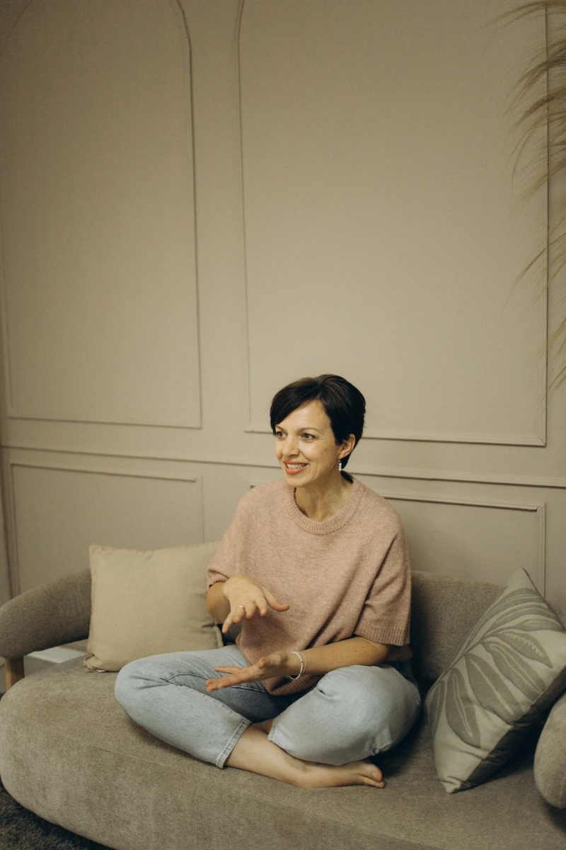 Anna Fasciani - freie Traurednerin -  sitzt barfuß und im Schneidersitz auf einem grauen Sofa mit Kissen und gestikuliert beim Gespräch.