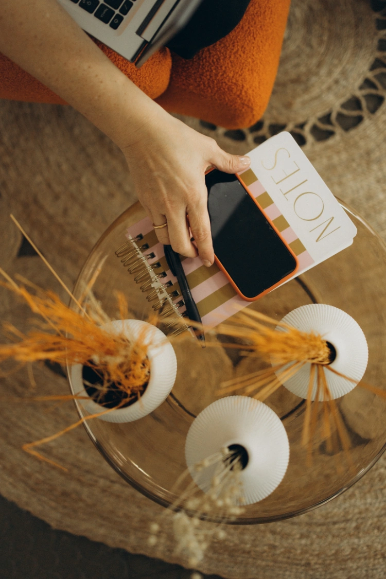 Hand hält Smartphone und Stift auf Notizblock mit Schriftzug 'NOTES' neben zwei weißen Vasen mit getrocknetem Gras auf Glas-Couchtisch.
