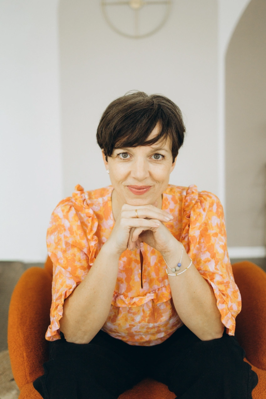 Anna Fasciani - freie Traurednerin -  mit kurzem dunkelbraunem Haar und orange gemustertem Oberteil, die lächelnd mit verschränkten Händen nach vorne sitzt.