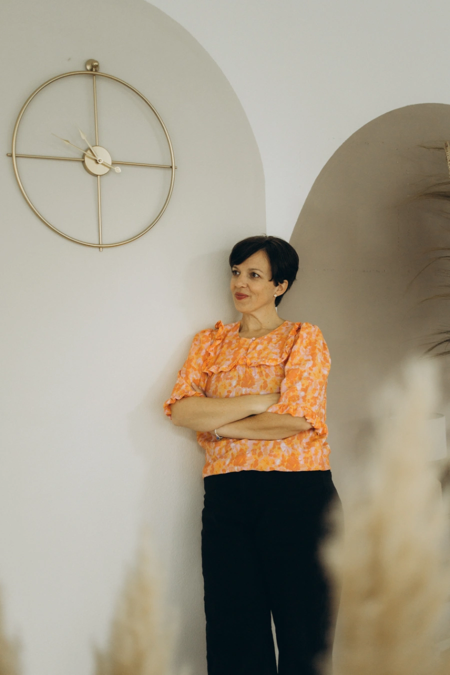 Anna Fasciani - freie Traurednerin -  in einer orange-weiß gemusterten Bluse und schwarzen Hosen lehnt mit verschränkten Armen an einer hellen Wand neben einer großen, runden goldfarbenen Uhr.