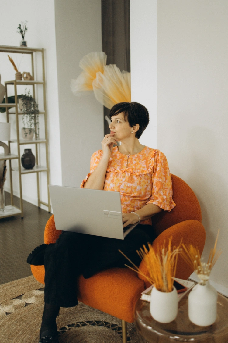 Anna Fasciani - freie Traurednerin -  in orangefarbenem Oberteil sitzt auf einem orangefarbenen Sessel mit einem Laptop auf dem Schoß und blickt nach links.
